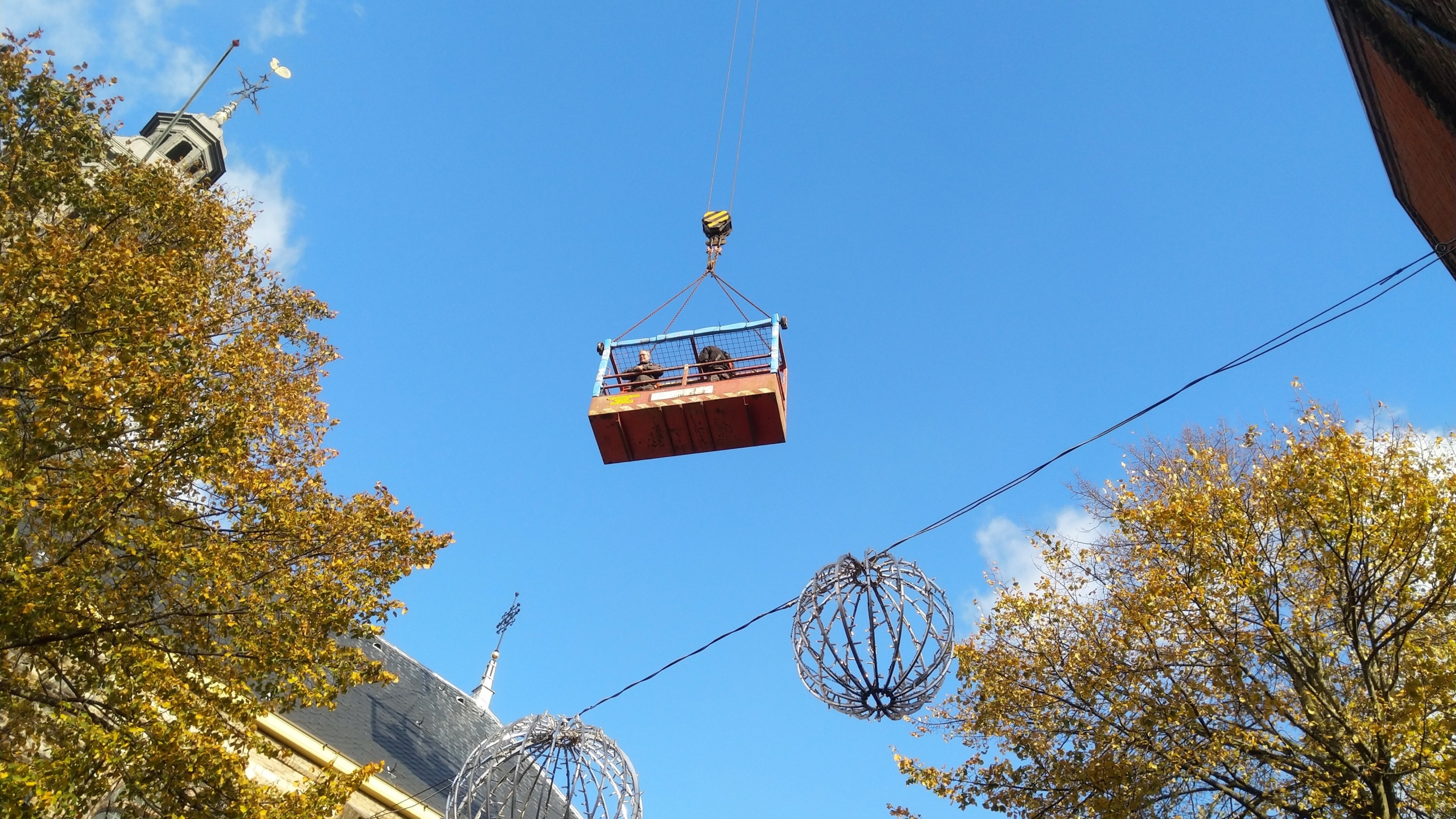 Mensen in een hangende metalen kooi boven een straat met herfstbomen en decoratieve bollen.