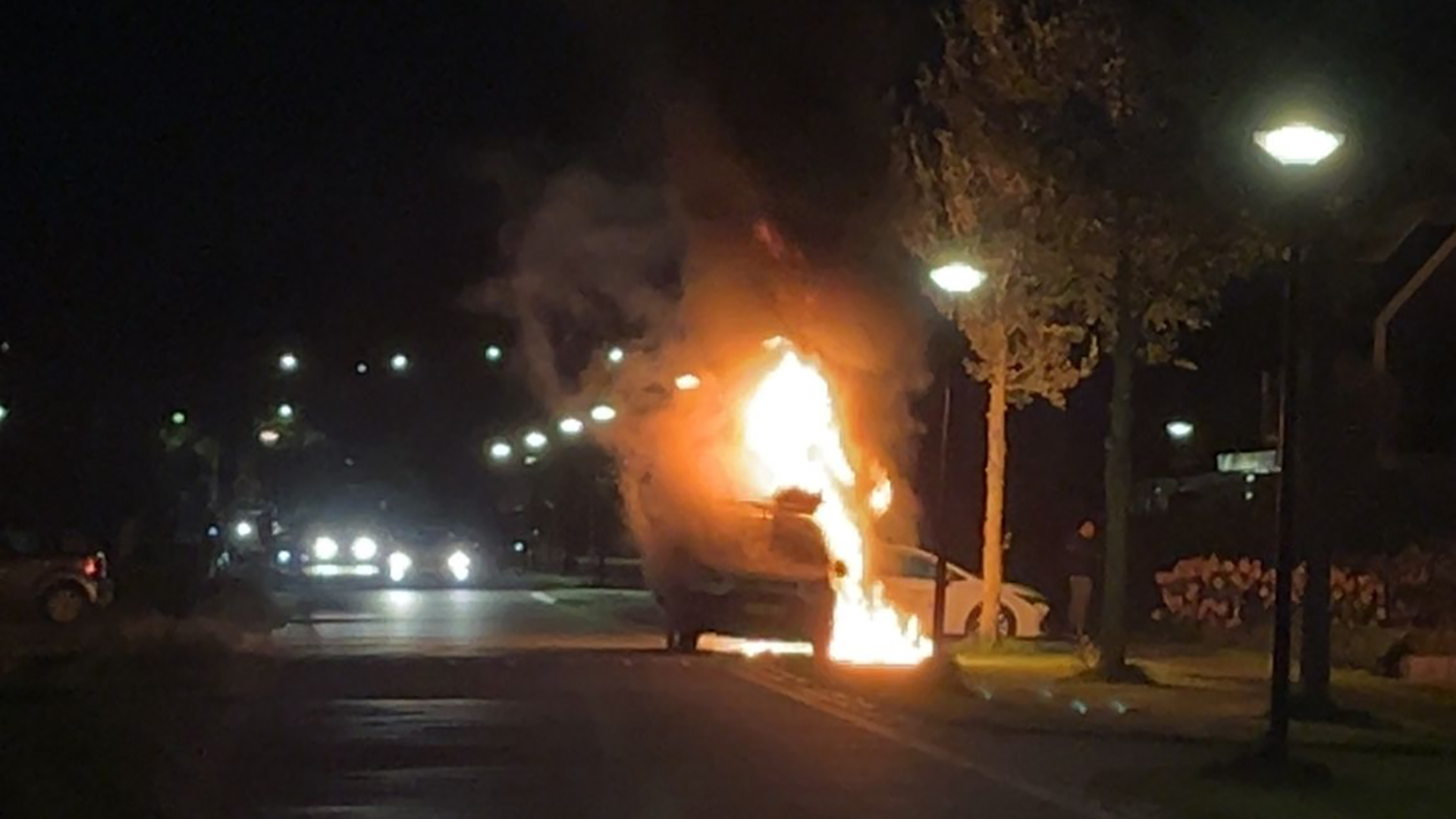 Auto in brand op straat 's nachts, met omliggende straatverlichting en rookwolken.