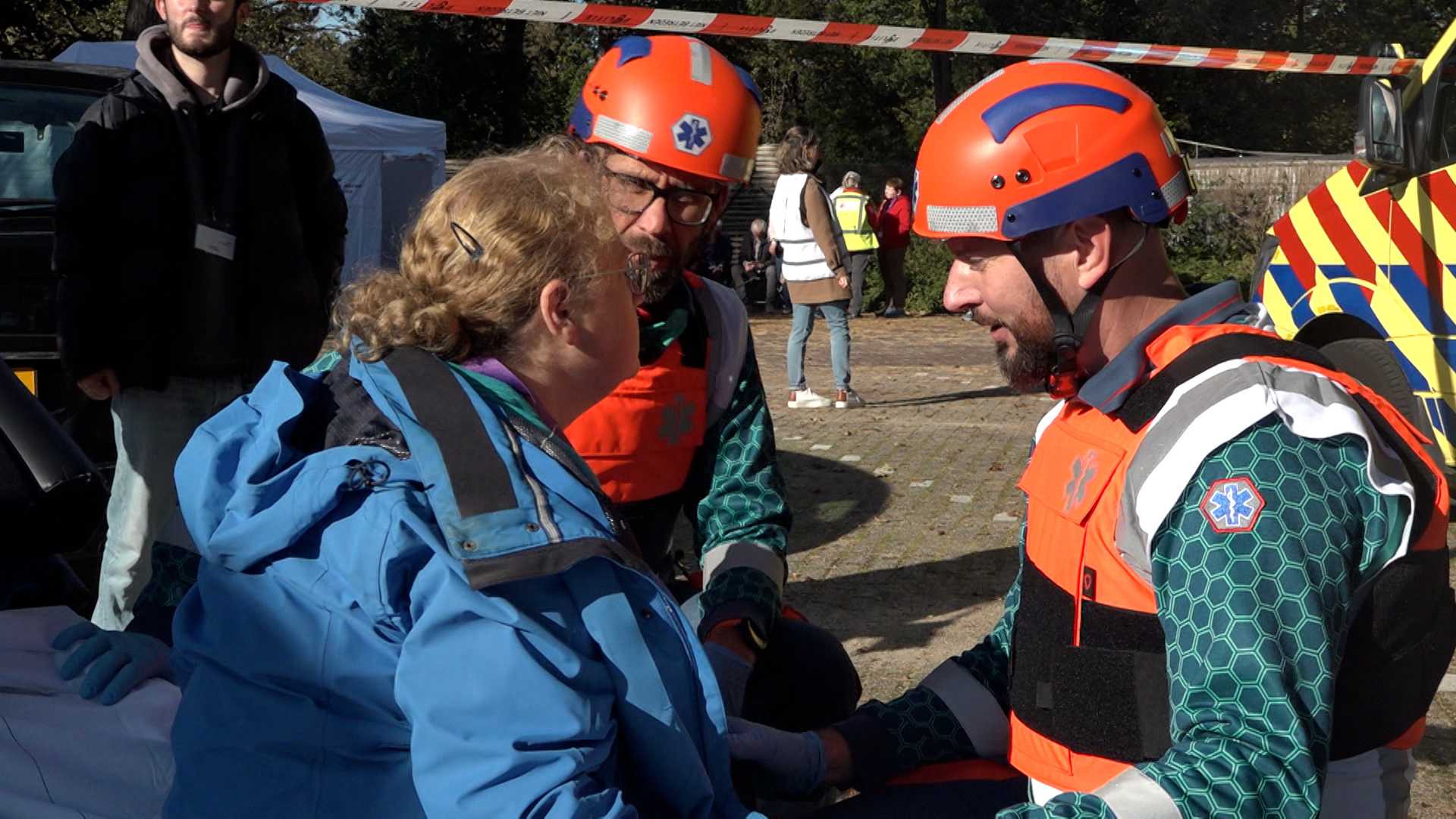 Hulpverleners met oranje helmen en reddingsvesten verlenen hulp aan een vrouw in blauwe jas.