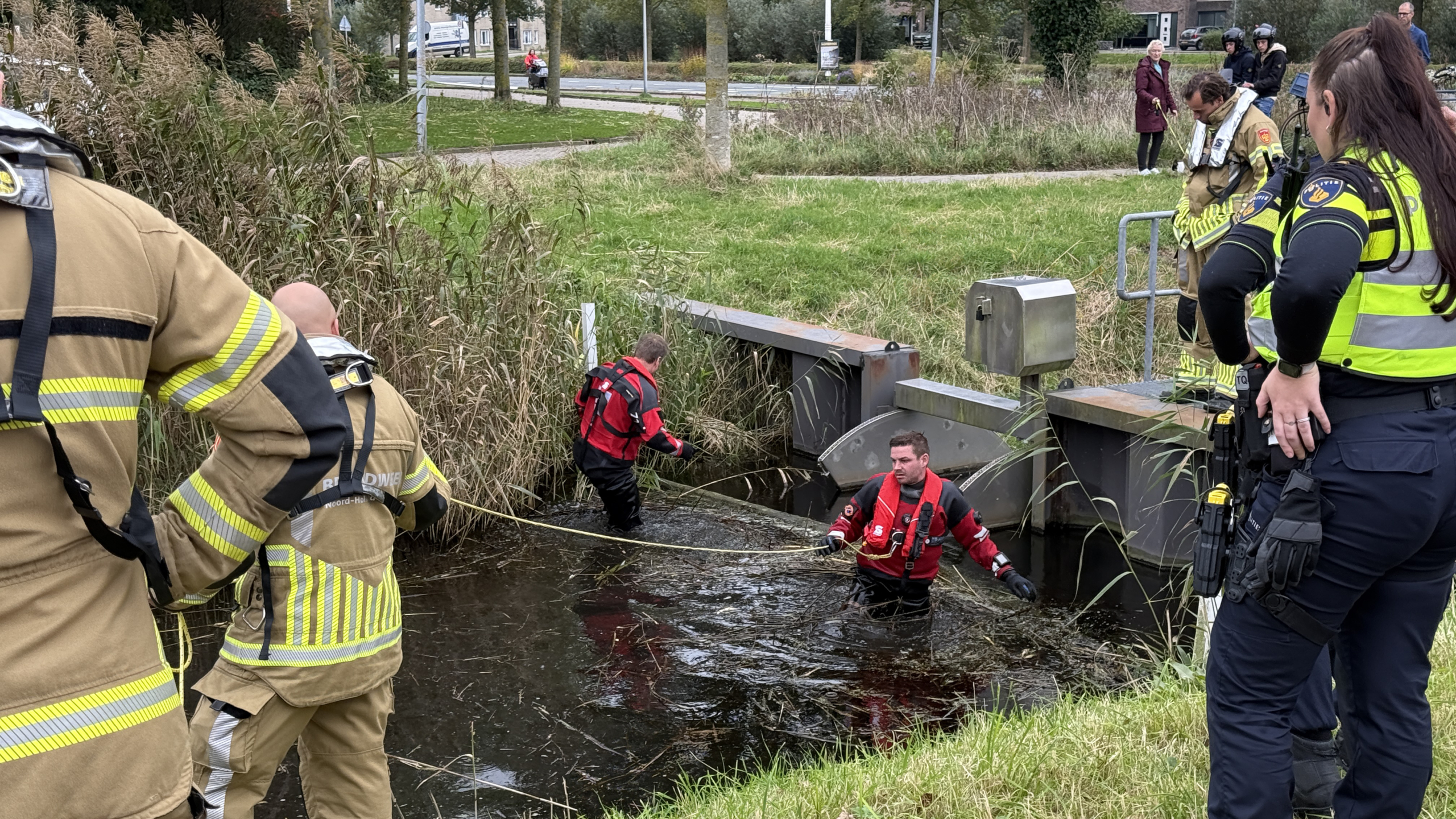 Hulpverleners in en bij een sloot, sommigen in waterreddingspakken, anderen in uniform op de oever.