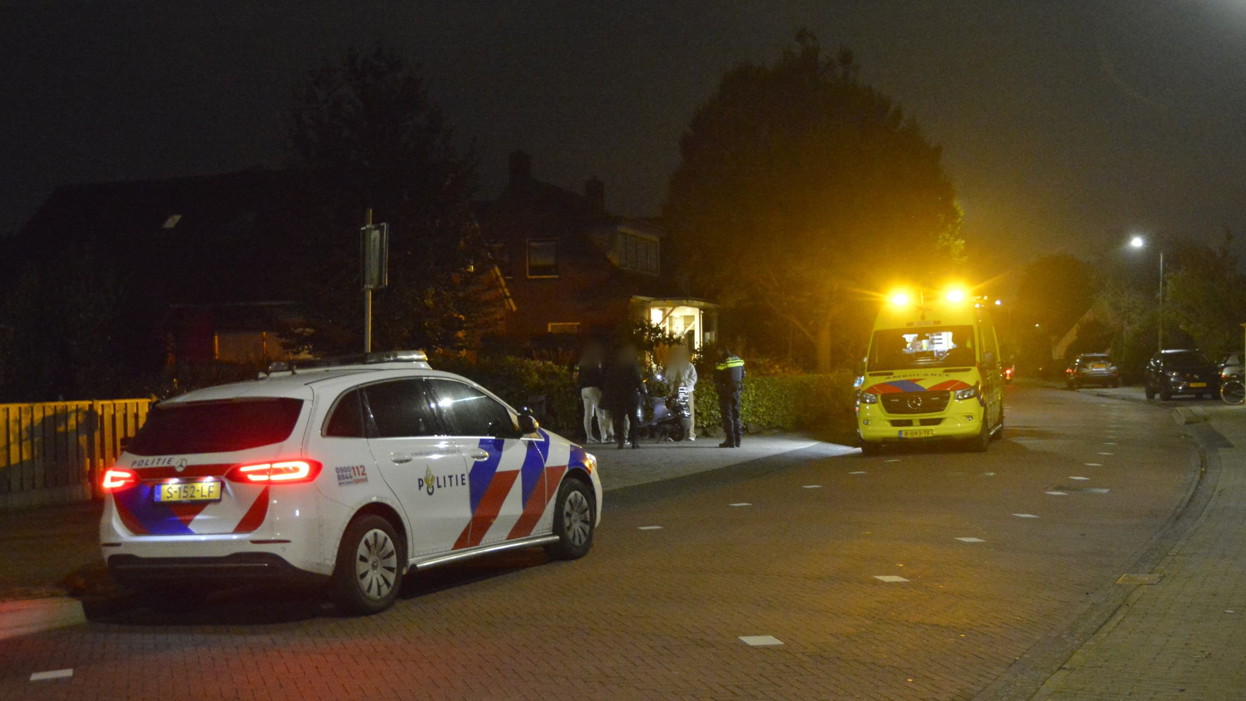 Politieauto en ambulance met flitslichten 's nachts op straat naast een huis, met mensen en een agent op het trottoir.