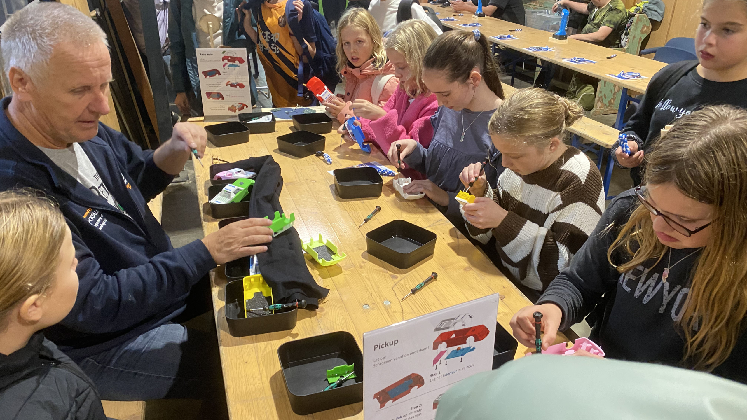 Kinderen en een volwassene nemen deel aan een workshop waarbij ze kleine plastic auto's in elkaar zetten aan een tafel.