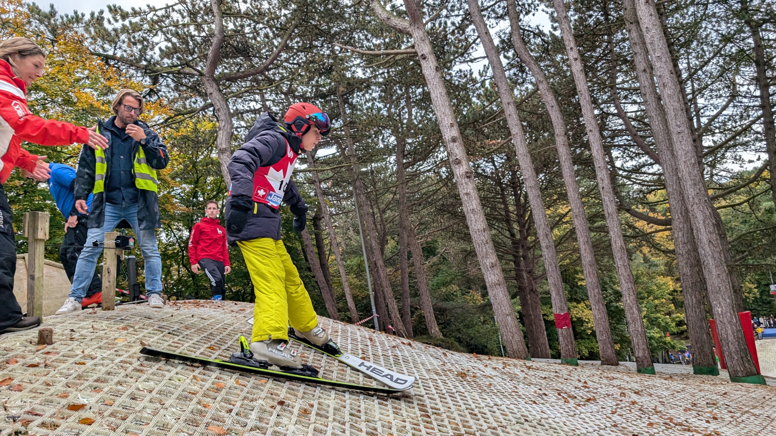 Een kind met een rode helm en gele skibroek op een kunstmatige skibaan, omringd door volwassenen en bomen.