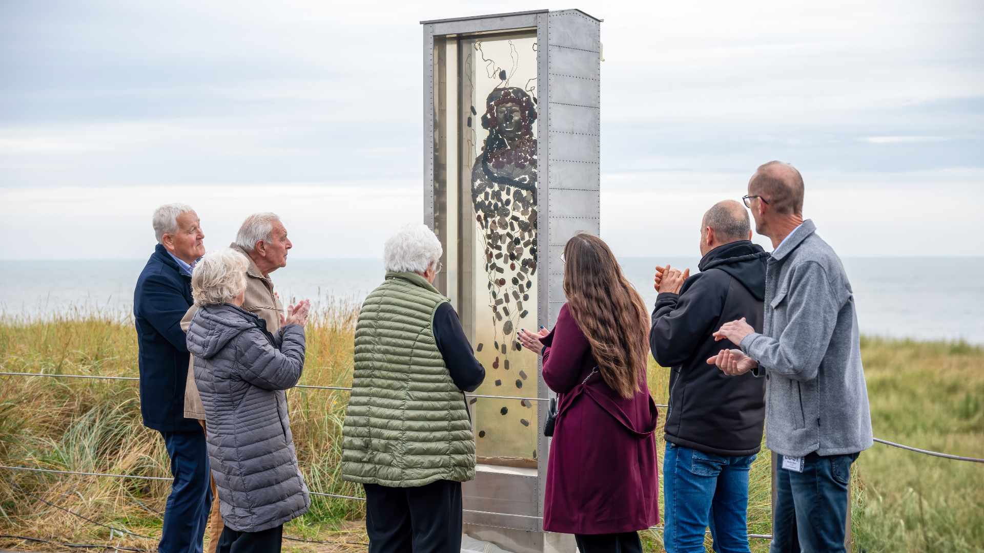 Een groep mensen kijkt naar een kunstwerk langs de kust.