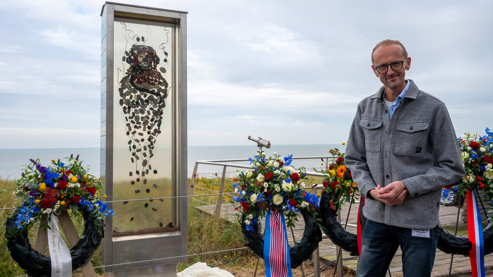 Man naast een monument met bloemstukken, met de zee op de achtergrond.