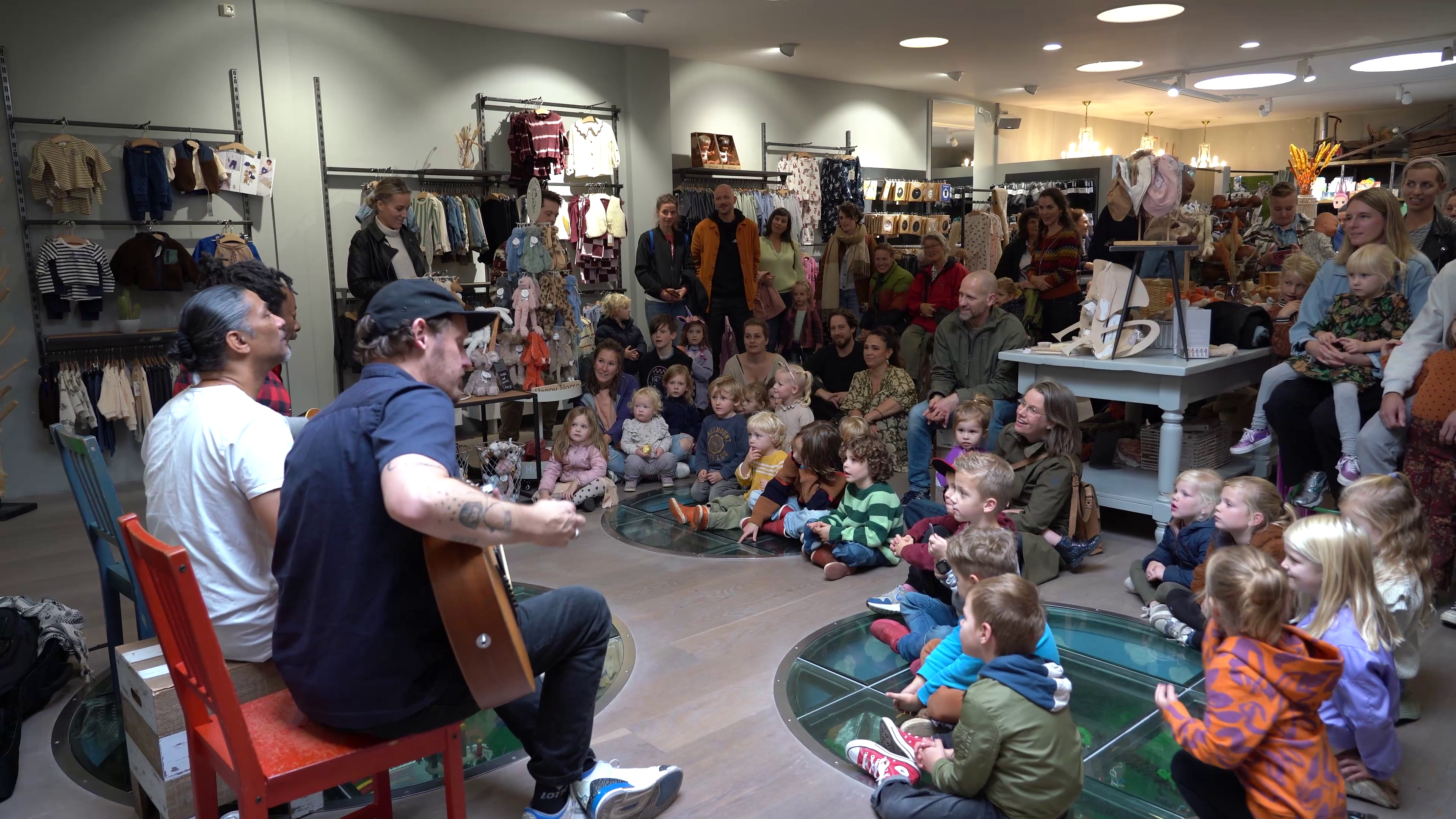 Muzikanten spelen gitaar voor een groep kinderen en volwassenen in een kinderkledingwinkel.