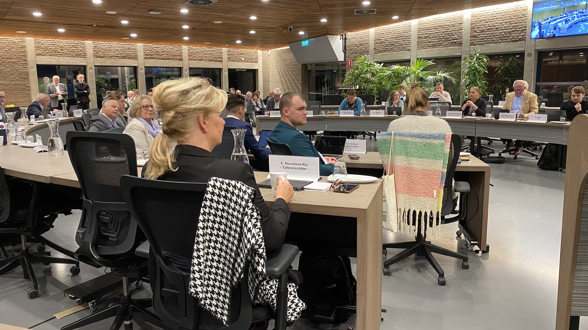 Vergadering in een moderne vergaderruimte met personen zittend aan tafels in een U-vorm.