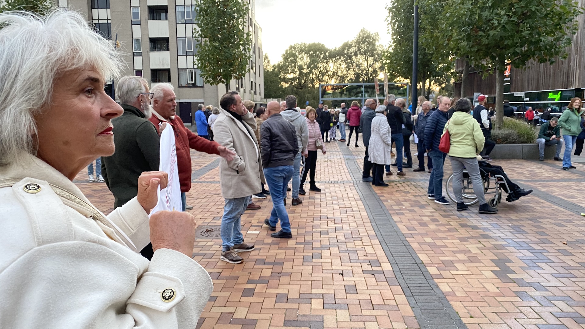 Mensen verzamelen zich buiten op een plein, enkele met protestborden, op een bewolkte dag.