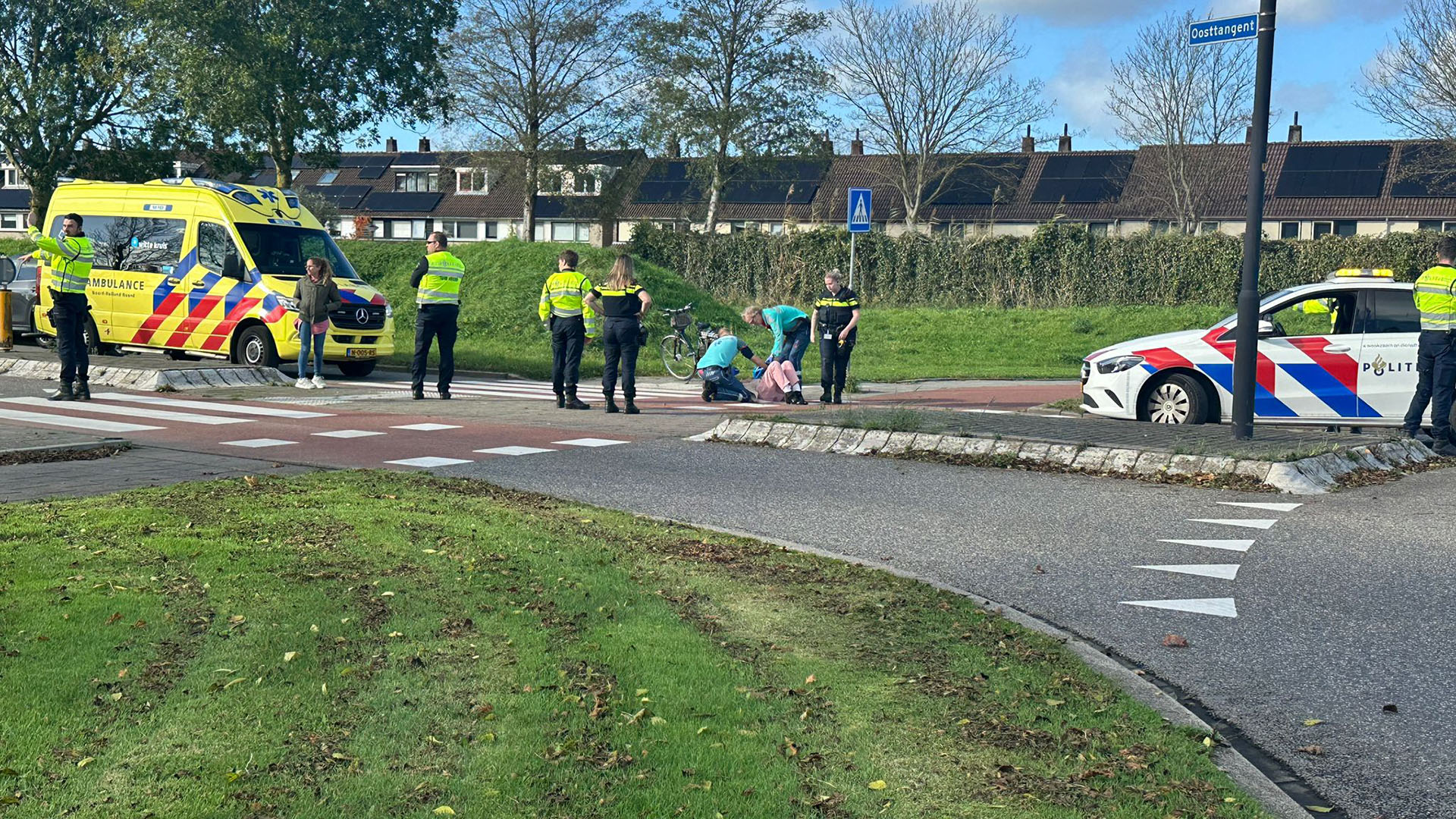 Een ongeval op een kruising met een ambulance, politieauto en meerdere politieagenten; hulpverleners zorgen voor een persoon op de grond bij een fietspad.