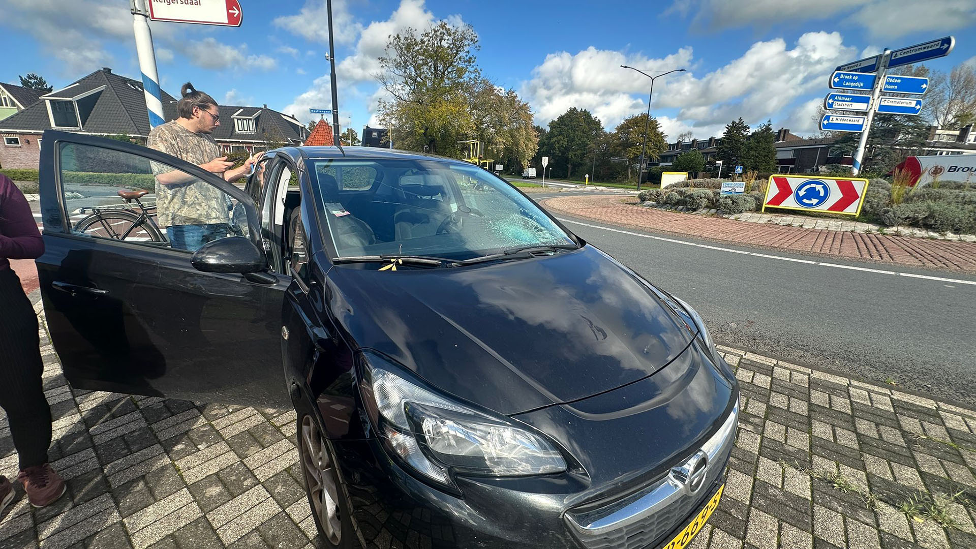 Een zwart voertuig met schade aan de voorruit staat geparkeerd bij een rotonde, met mensen die naast het geopende portier staan.