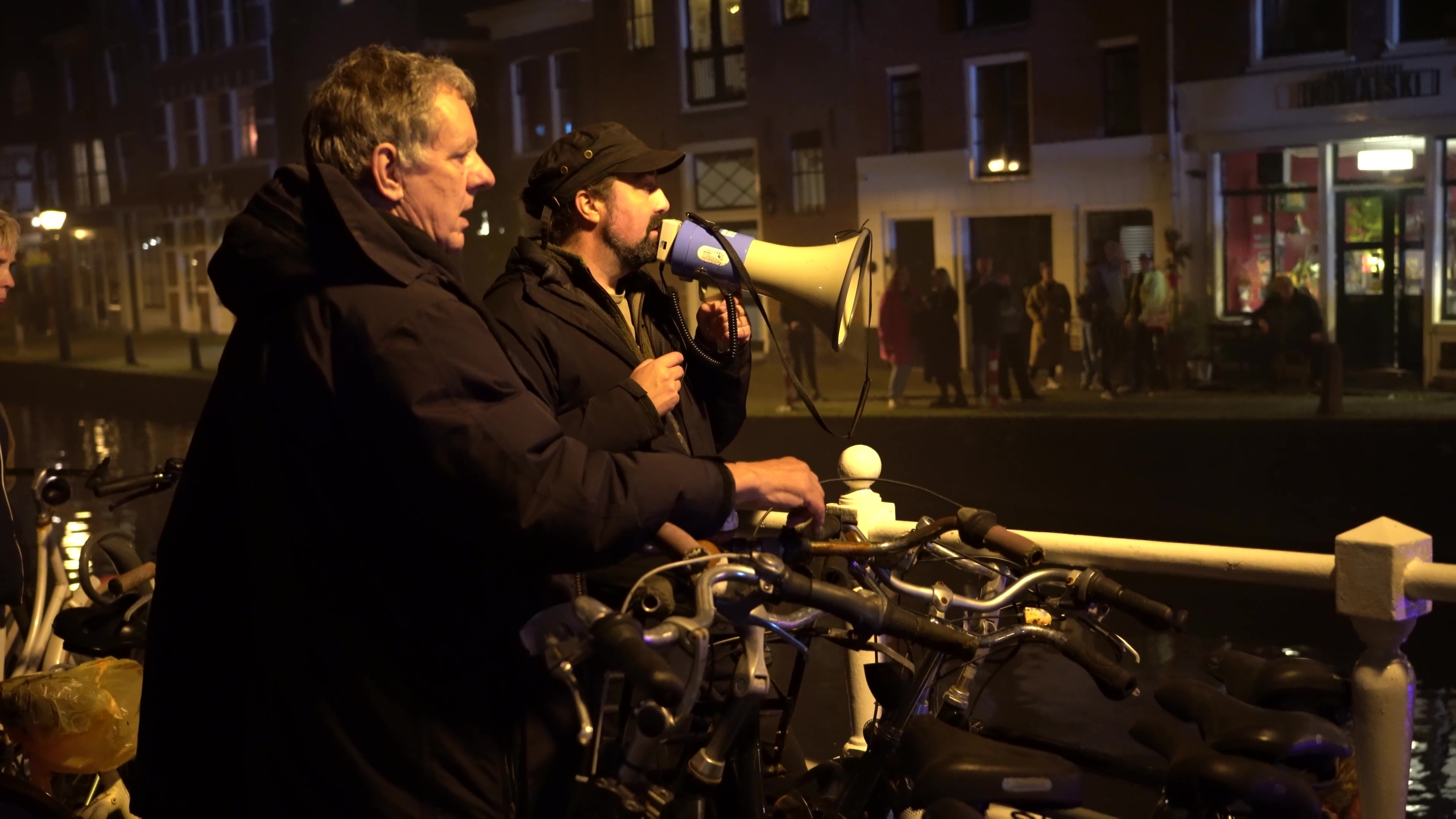 Twee mannen dragen donkere jassen, waarvan één in een megafoon spreekt, op een brug bij nacht, met fietsen op de voorgrond en mensen op de achtergrond.