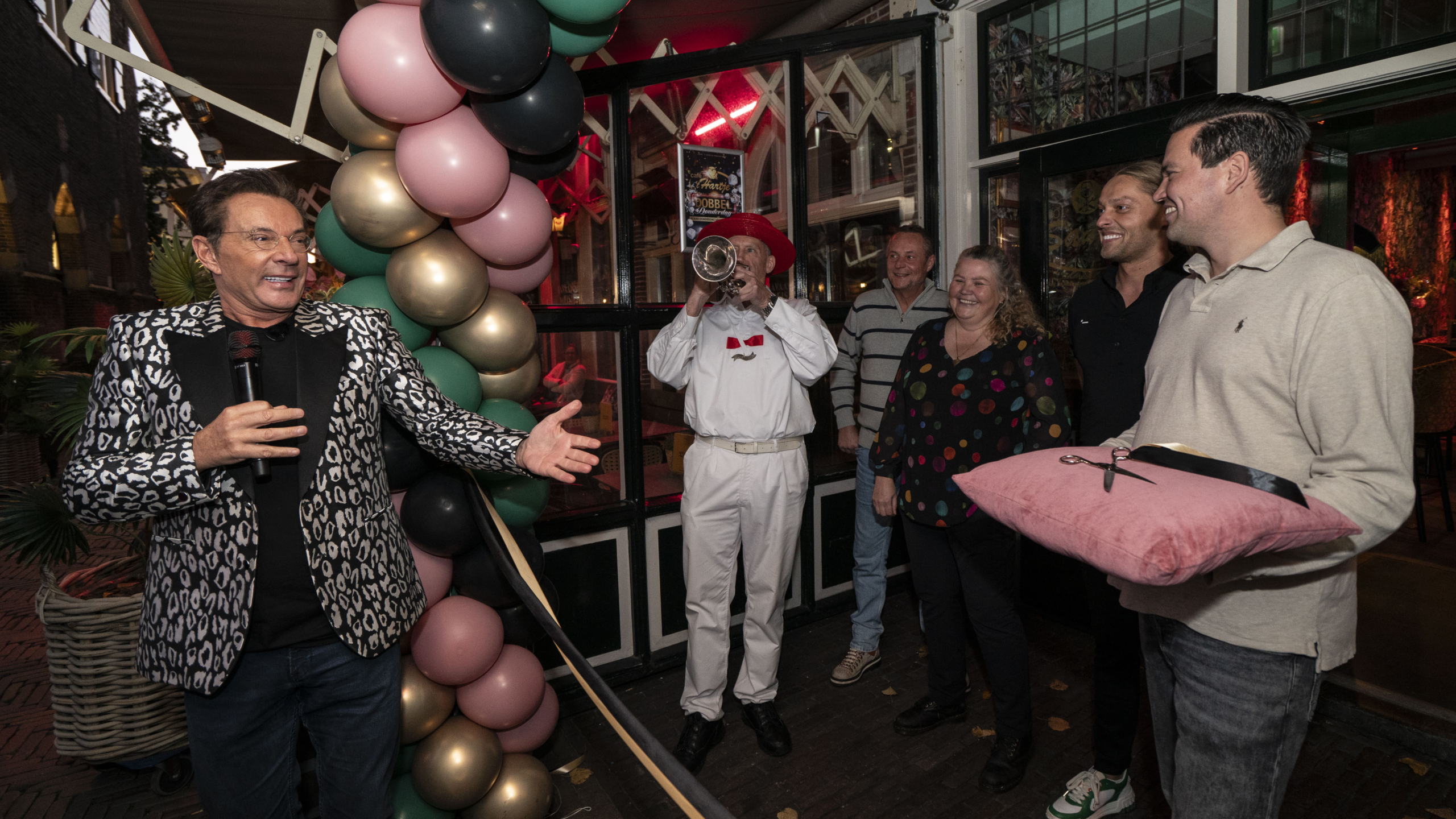 Man met microfoon in feestelijke jas naast ballonboog, muzikant met trompet en groep lachende mensen. Een man houdt een roze kussen met schaar vast.