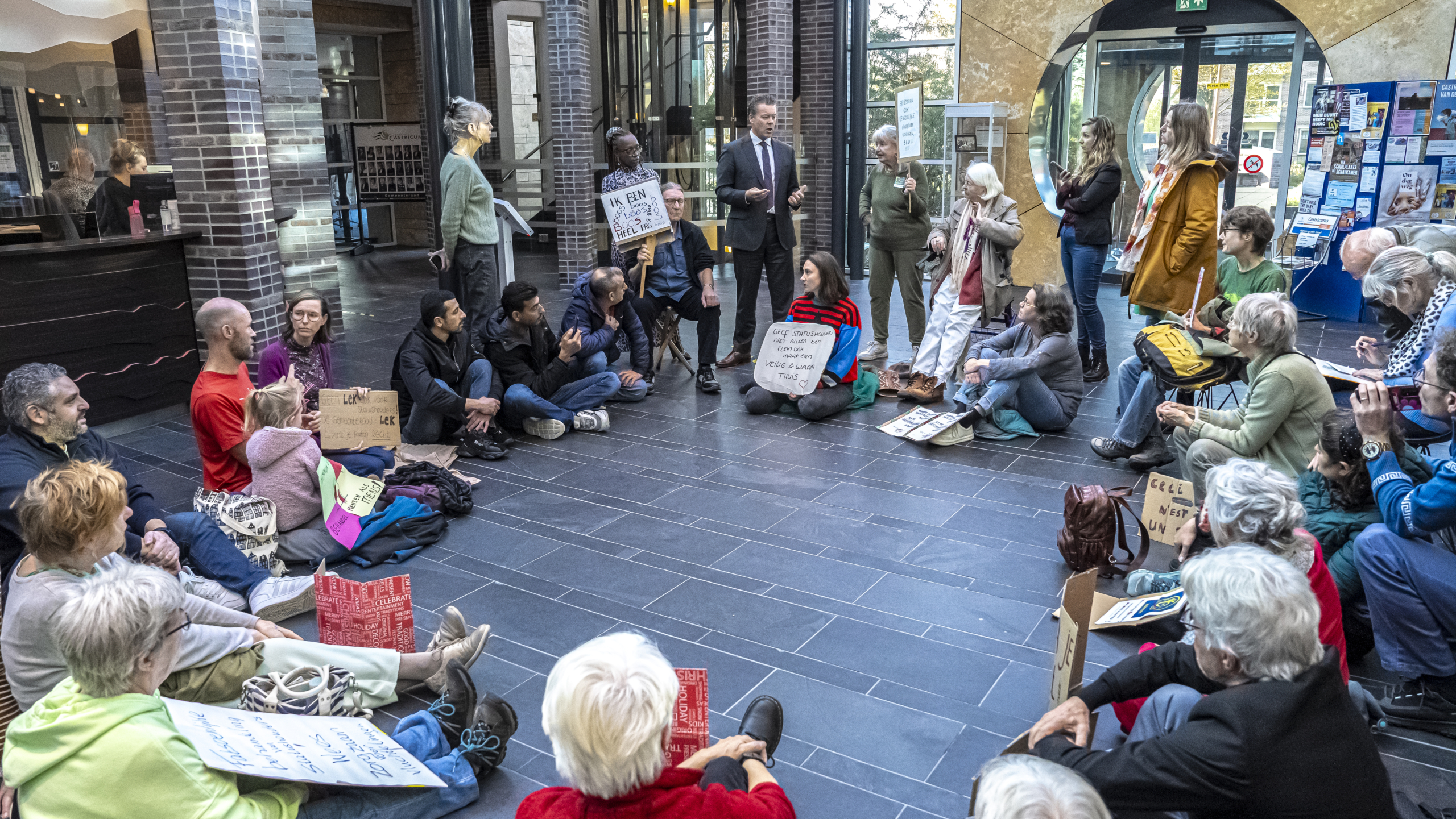 Mensen zitten op de grond in een openbare ruimte en houden protestborden vast terwijl iemand hen toespreekt.