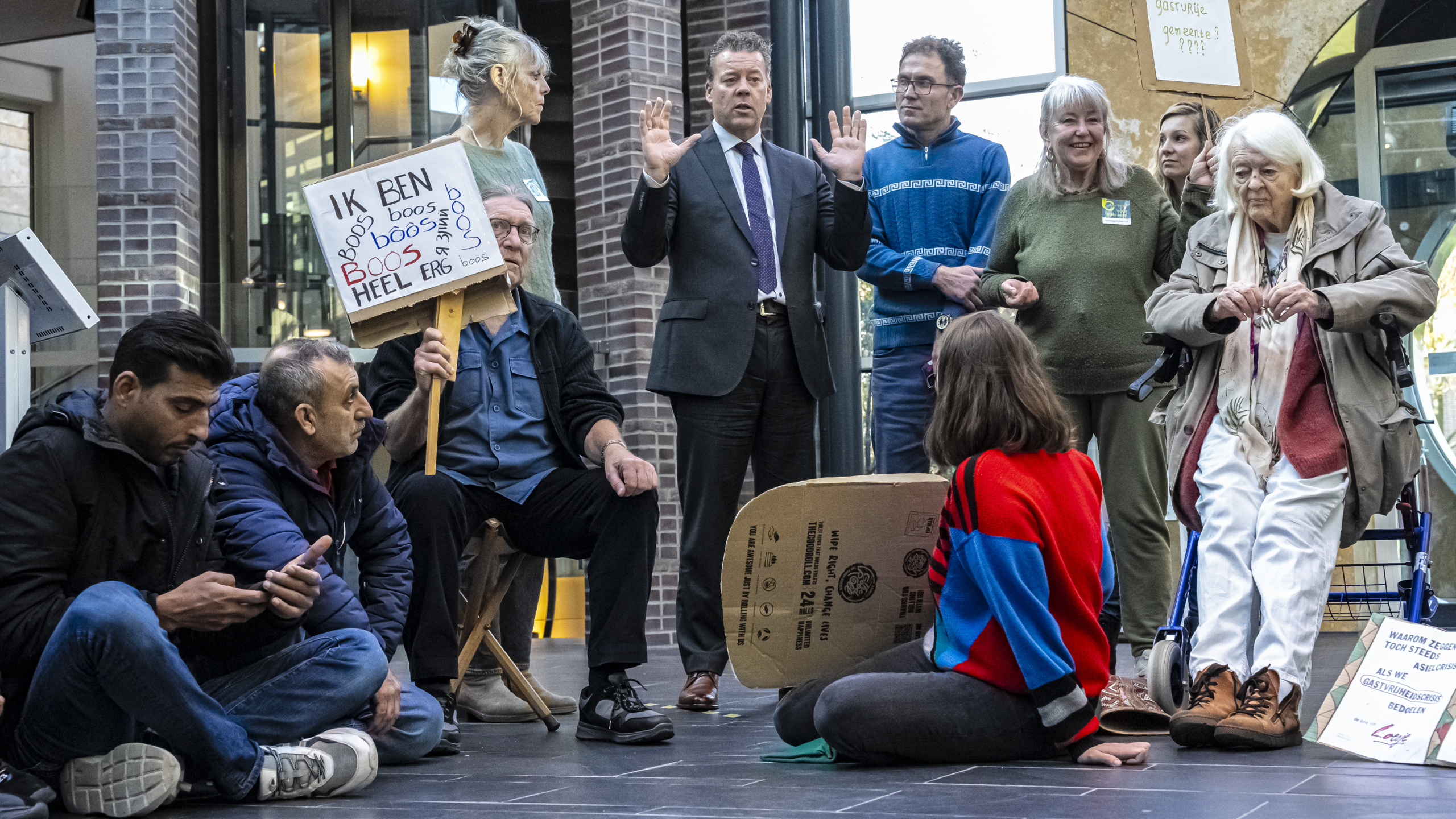 Een groep mensen, waarvan sommigen zitten en anderen staan, met protestborden tijdens een demonstratie in een binnenruimte. Eén bord met tekst "Ik ben heel erg boos".