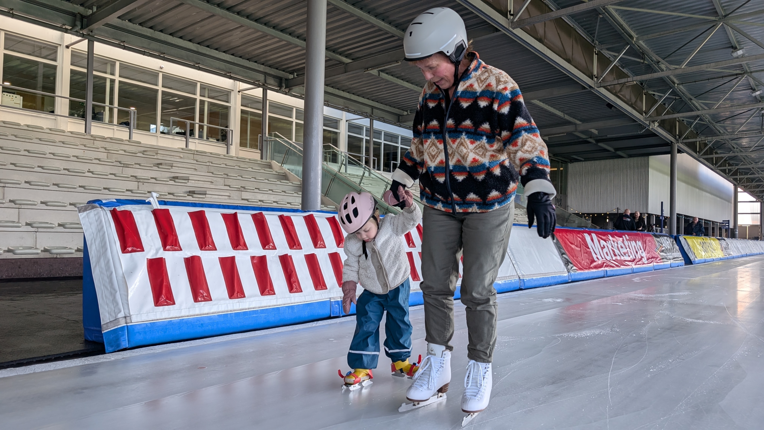 Een kind en volwassene schaatsen samen op een ijsbaan, beiden dragen helmen.