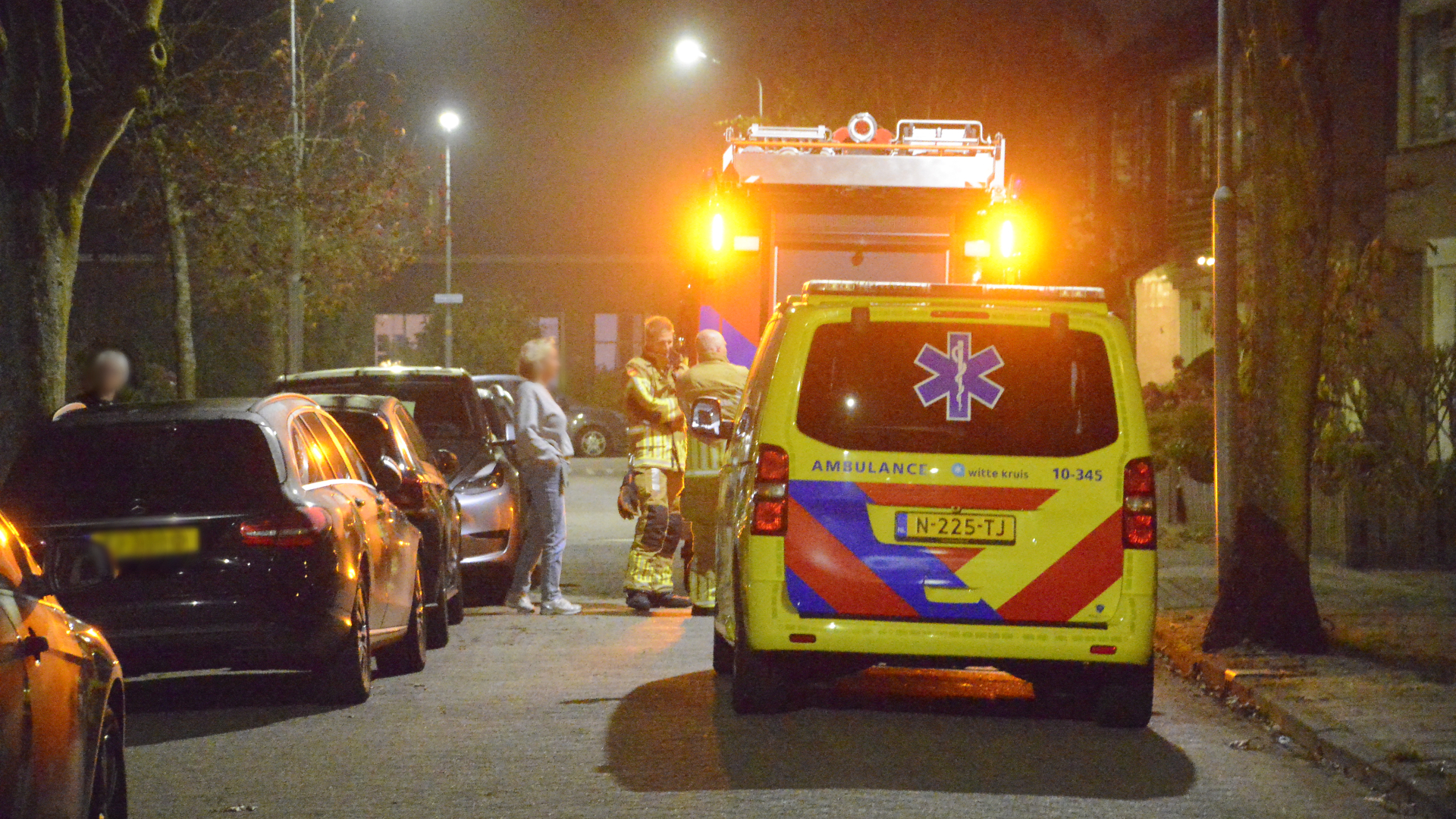 Een ambulance en brandweerwagen met zwaailichten staan 's nachts geparkeerd op een straat, met hulpverleners die overleggen naast de voertuigen.