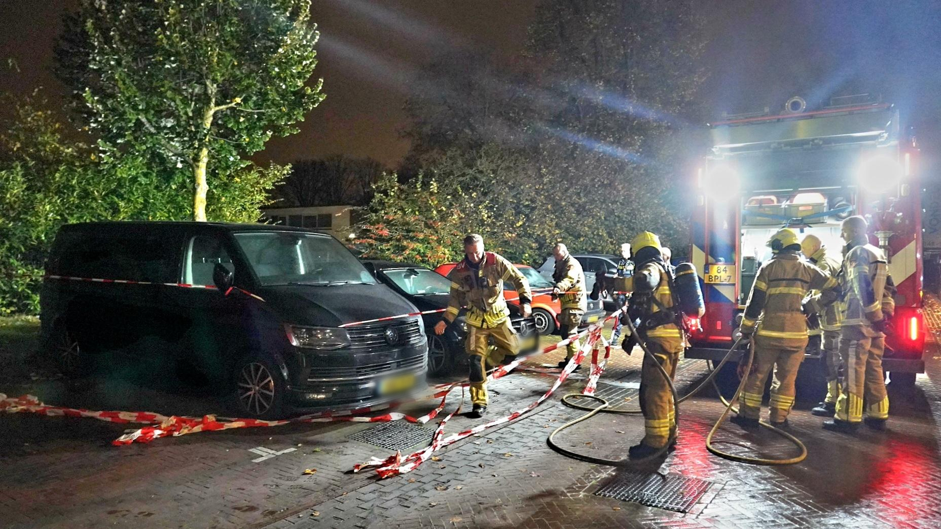 Brandweerlieden bij nachtelijke inzet naast geparkeerde voertuigen, met afzetlint en brandweervoertuig.