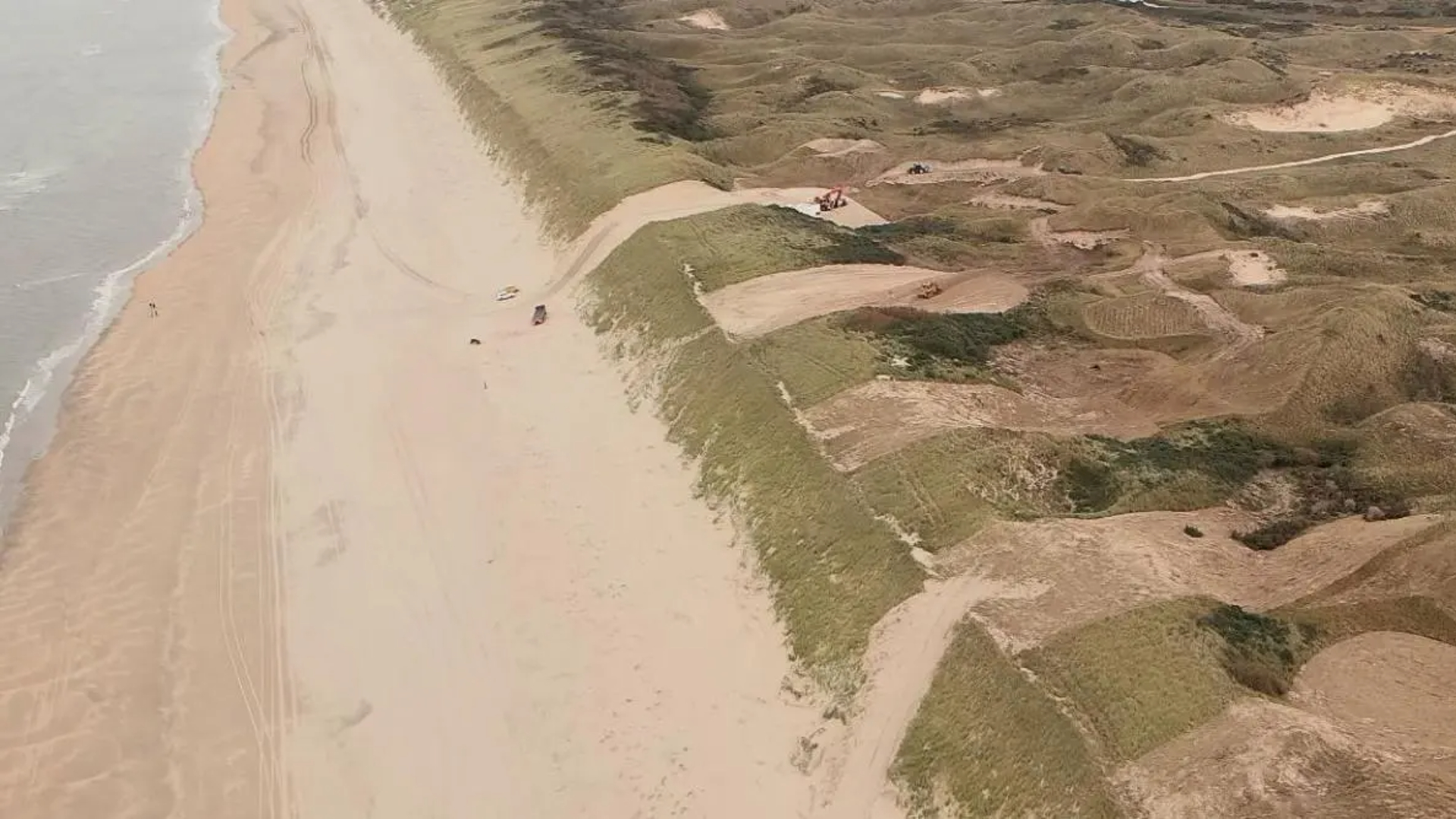 Luchtfoto van een strand met duinen en enkele voertuigen op het zand.