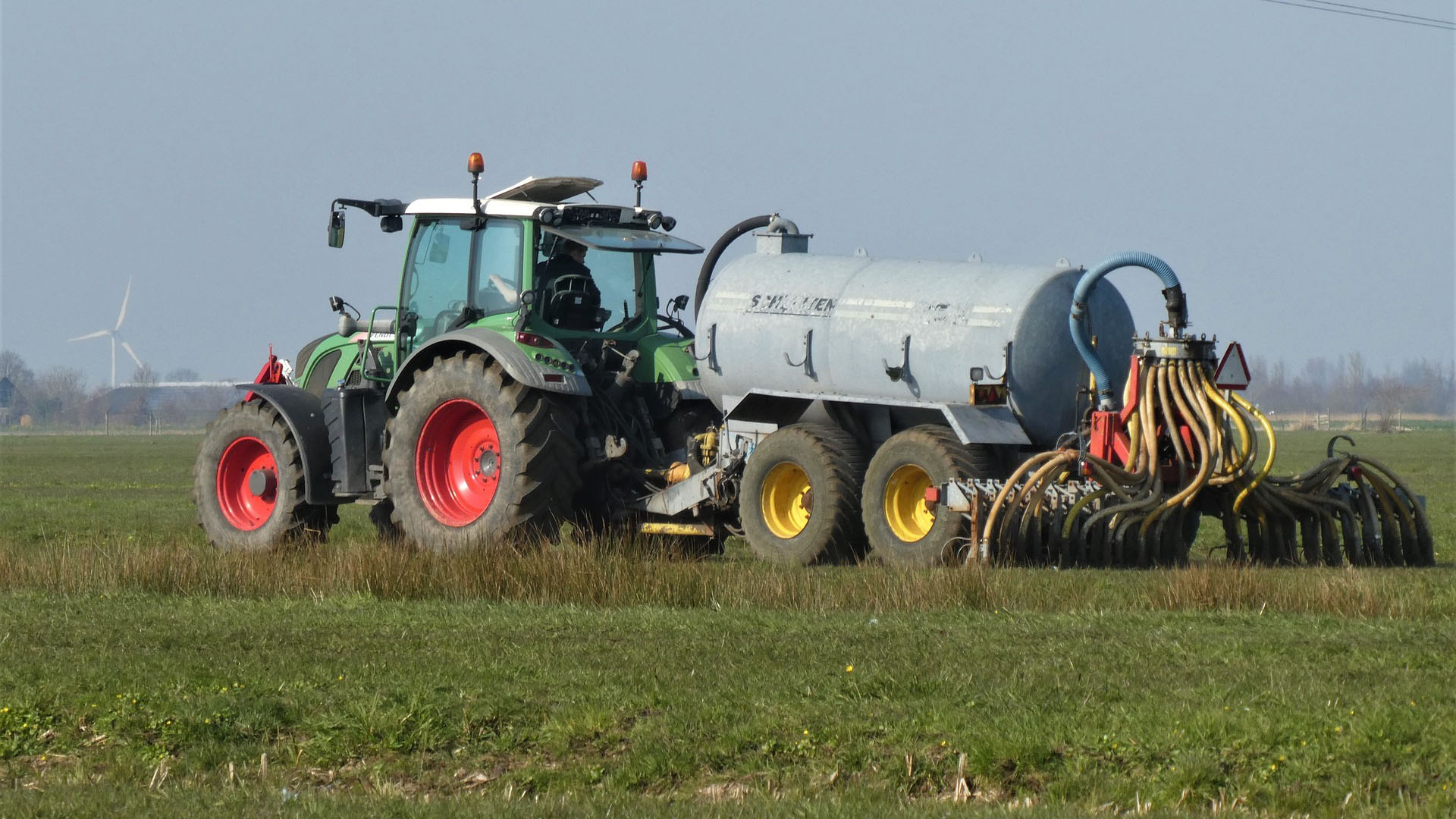 Tractor met mesttank op een grasveld met windmolen op de achtergrond.