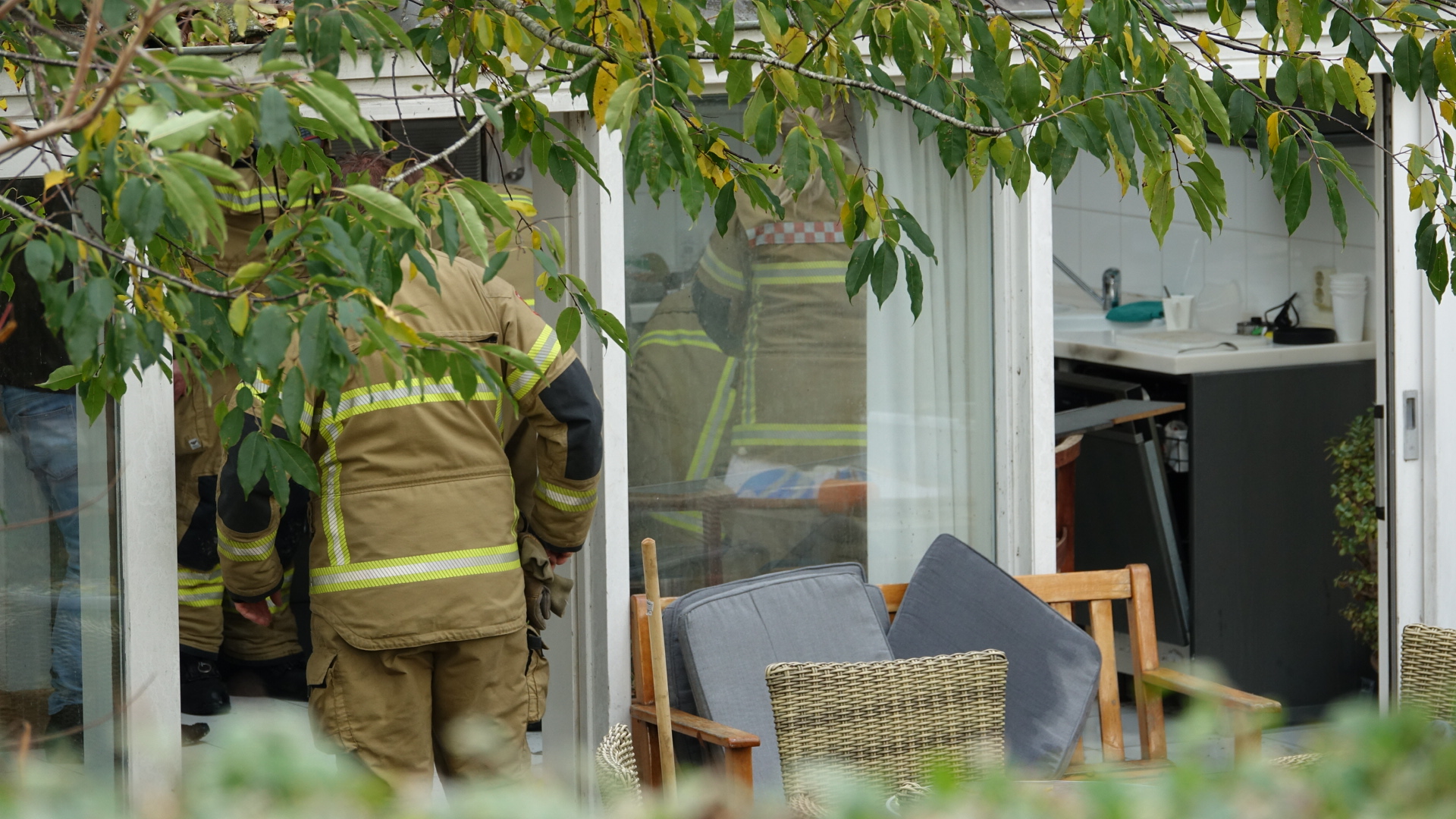 Brandweerman in beschermende kleding staat in deuropening van huis met zicht op binnenruimte en planten.