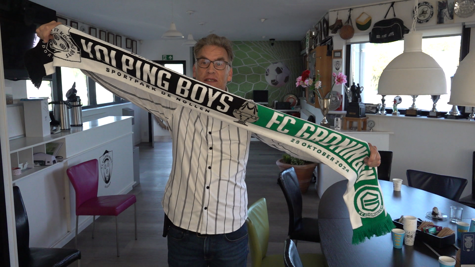 Man houdt een sjaal omhoog in een kamer met trofeeën en voetbaldecoraties, met de tekst "Kolping Boys" en "FC Groningen" erop.