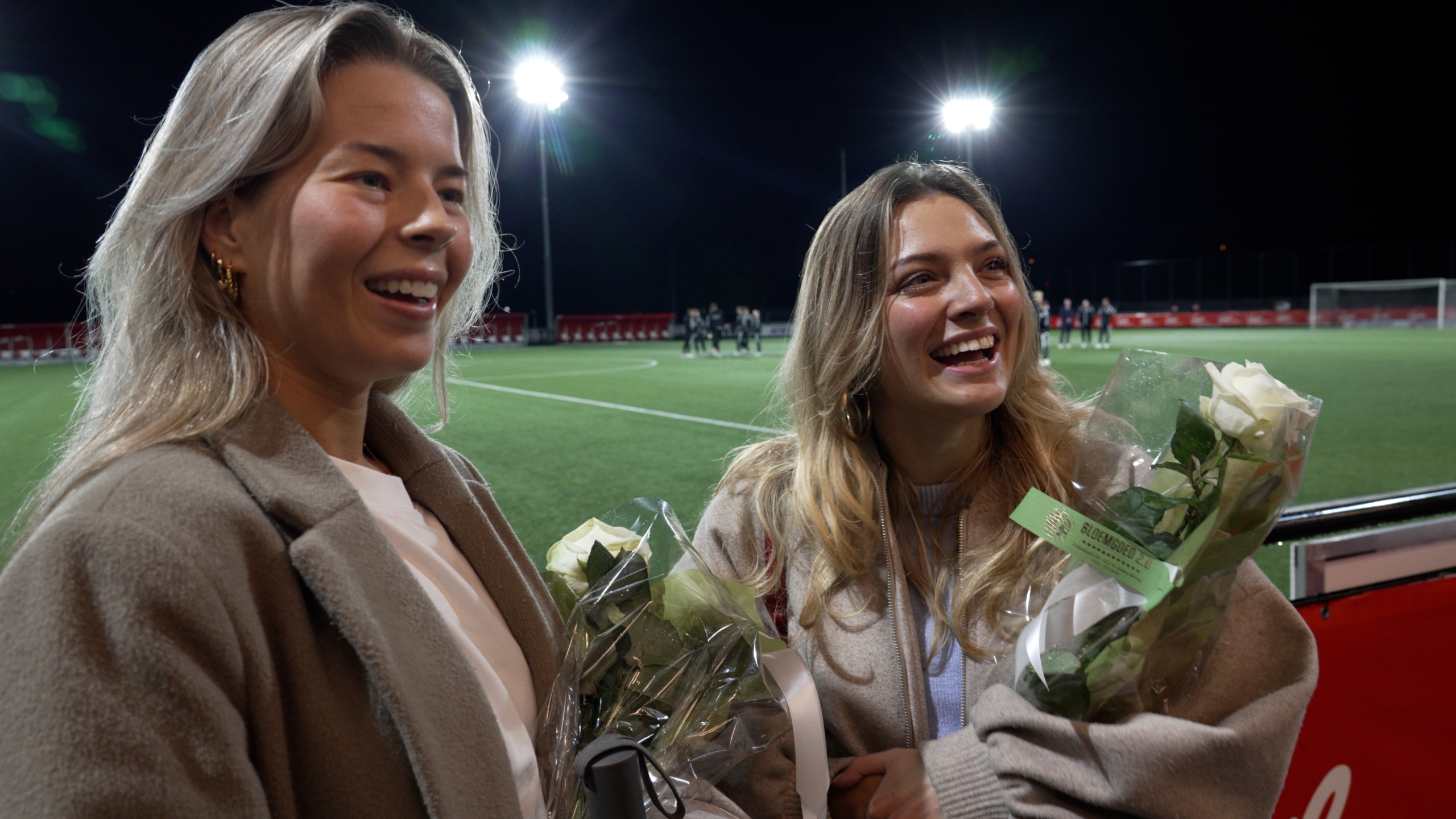 Twee jonge vrouwen lachen en houden bloemen vast op een verlicht voetbalveld.