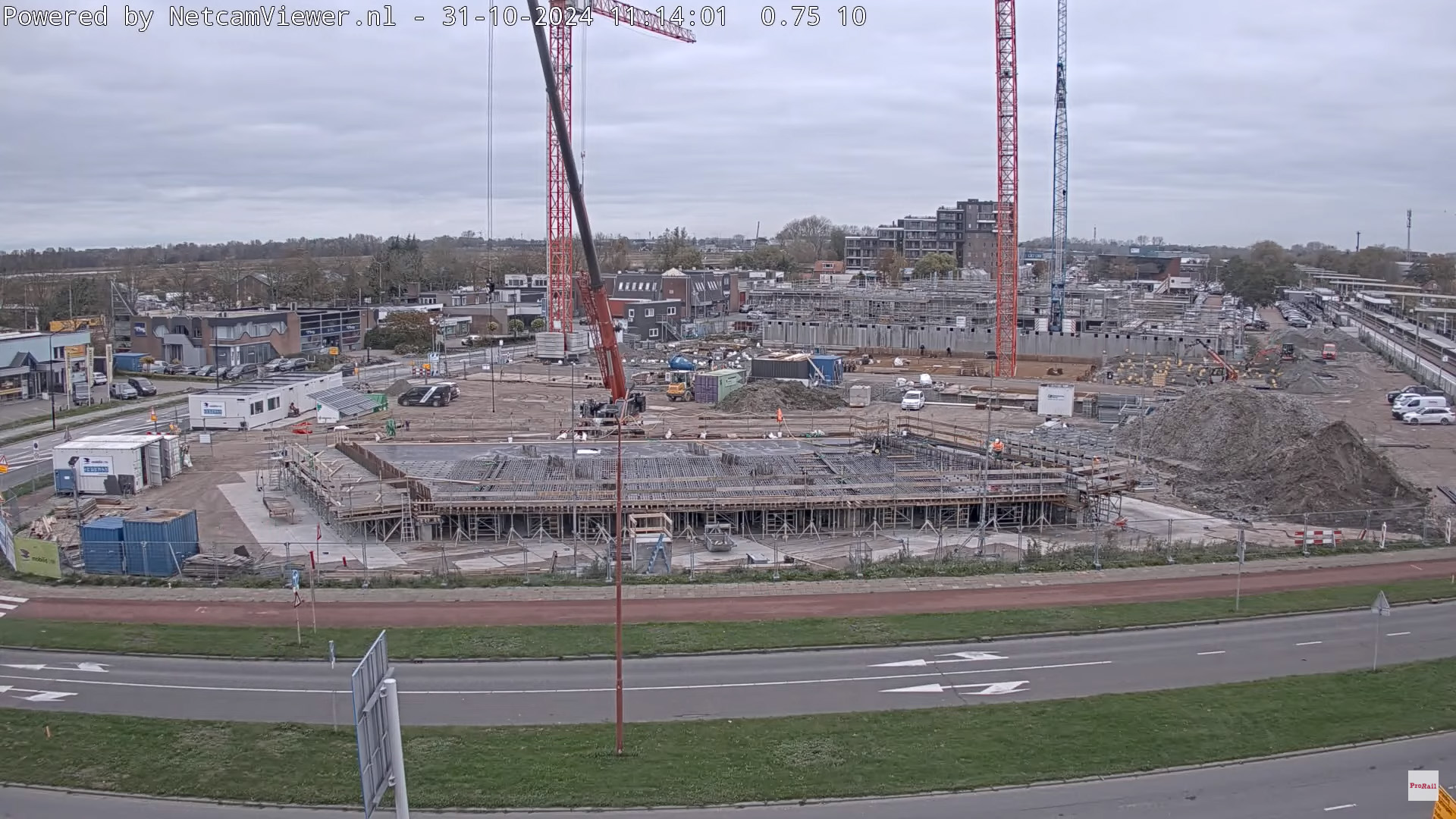 Bouwplaats met hijskranen, betonconstructies en bouwmaterialen, omringd door wegen en gebouwen.