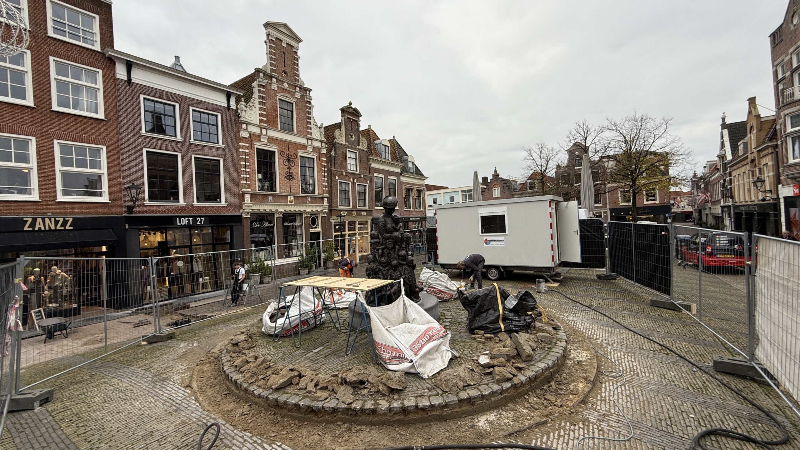 Straat met historische gebouwen en winkels, middelpunt met een omheinde fontein in aanbouw, bouwmaterialen en arbeiders aanwezig.