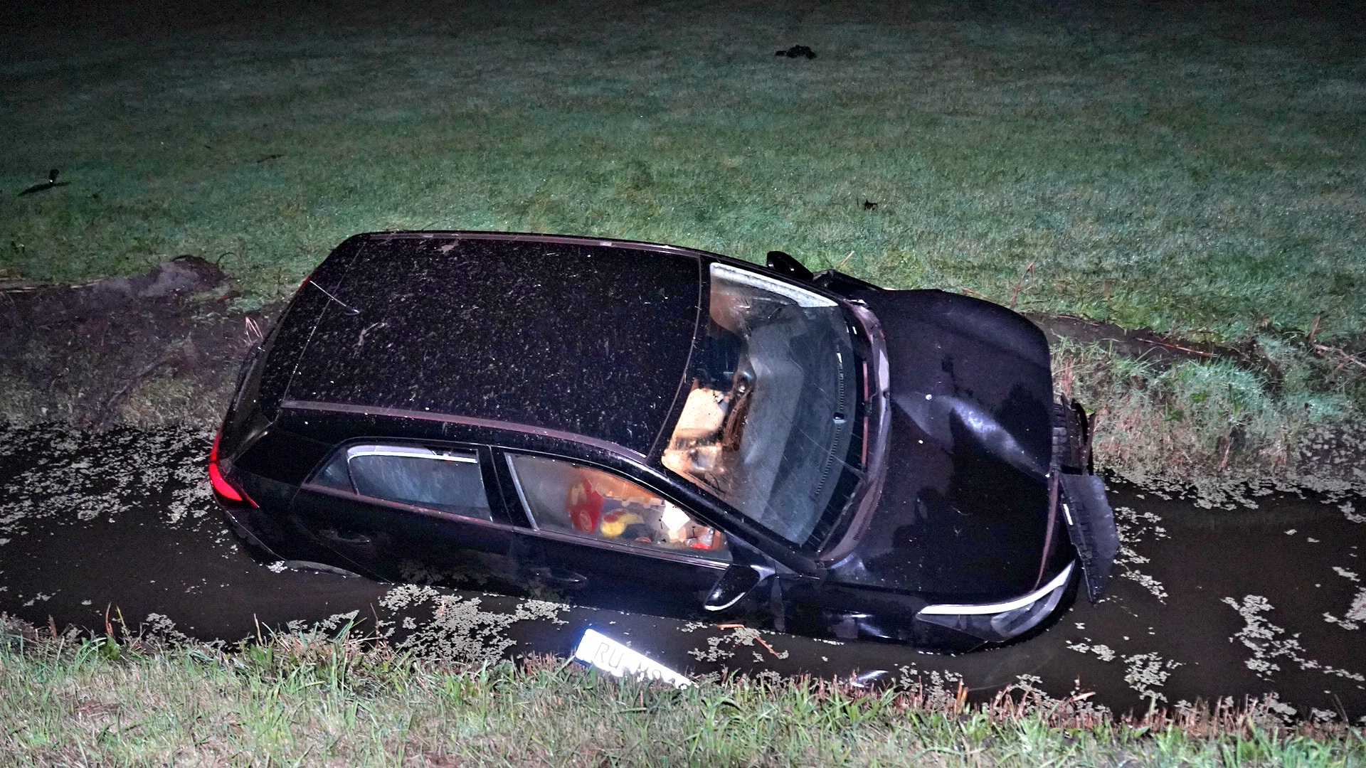 Zwarte auto in een greppel met water, met schade aan de voorkant, omgeven door gras in de nacht.