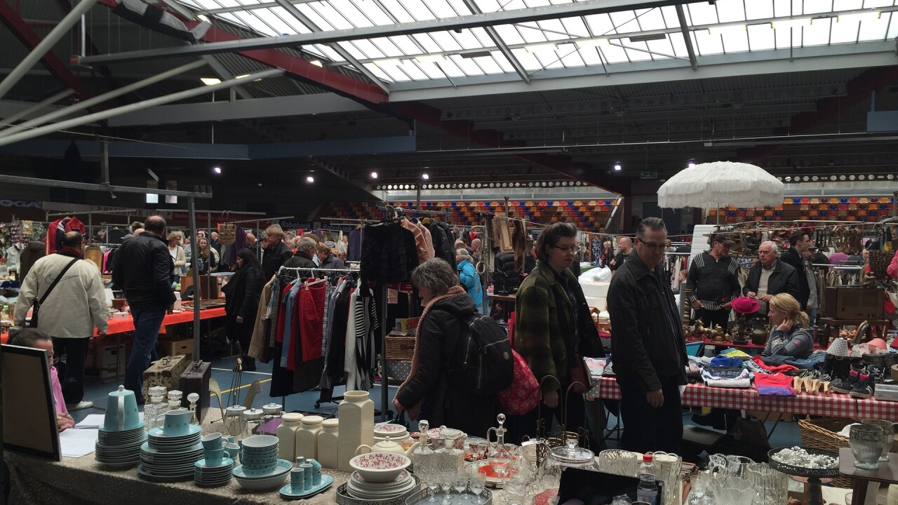 Mensen winkelen en bekijken artikelen op een druk bezochte rommelmarkt in een grote overdekte ruimte.