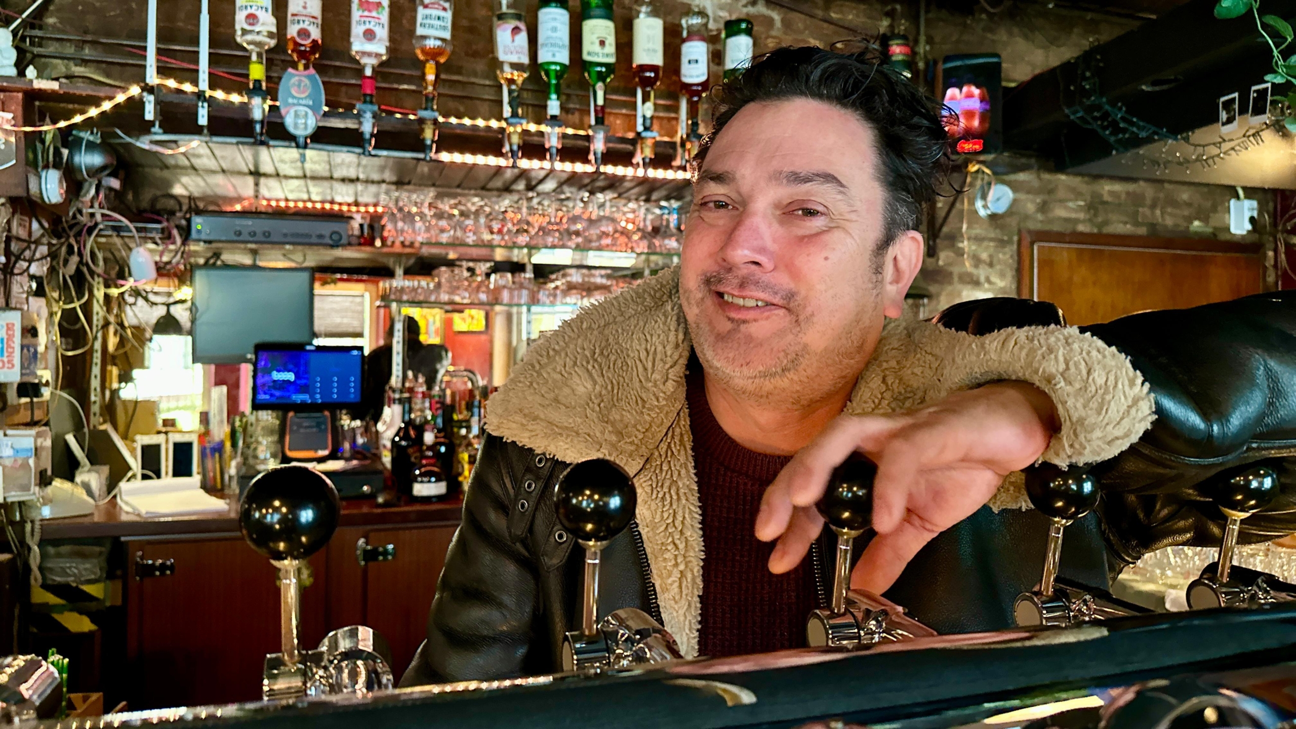 Man leunt glimlachend op de bar in een café met flessen drank en glazen op de achtergrond.