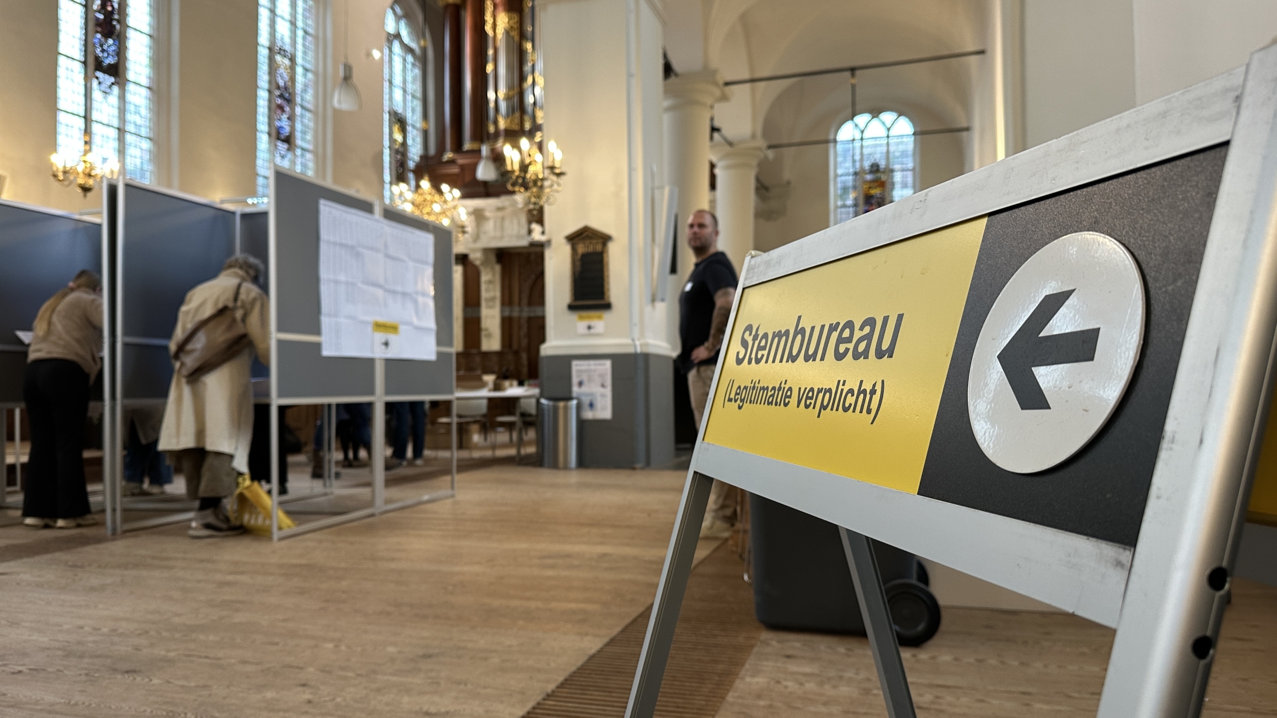 Bord met tekst "Stembureau, legitimatie verplicht" in een stemlokaal met mensen die stemmen.