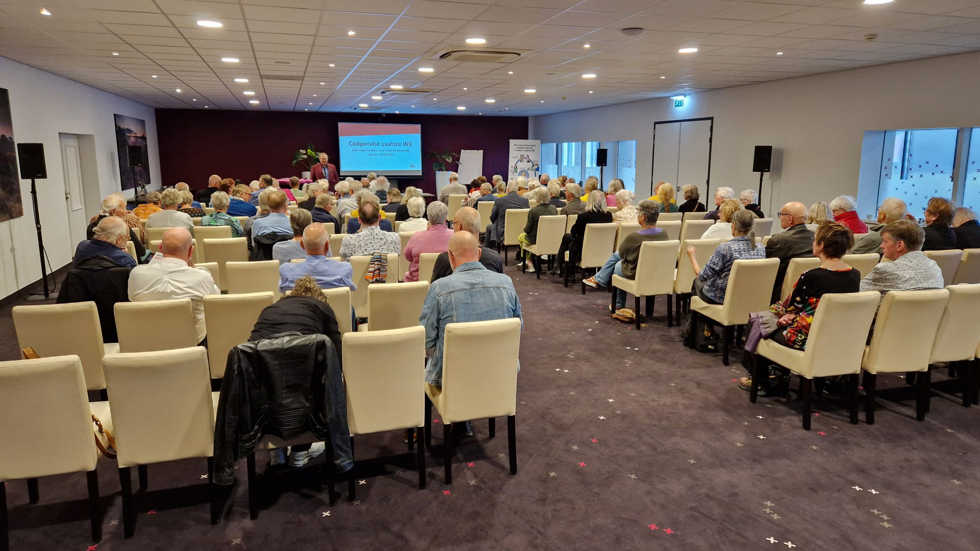 Een zaal vol mensen die een presentatie bekijken met een scherm waarop "Coöperatie Laatste Wil" staat.