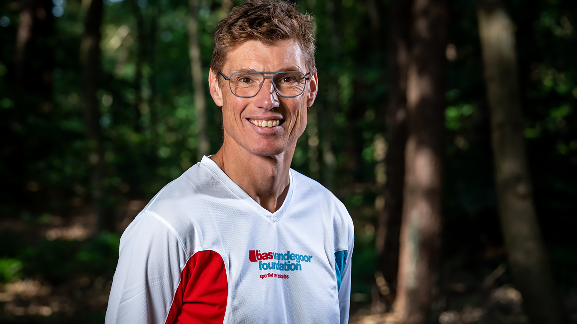 Man met bril en witte sportshirt met logo van 'Bas van de Goor Foundation', lachend in een bosrijke omgeving.