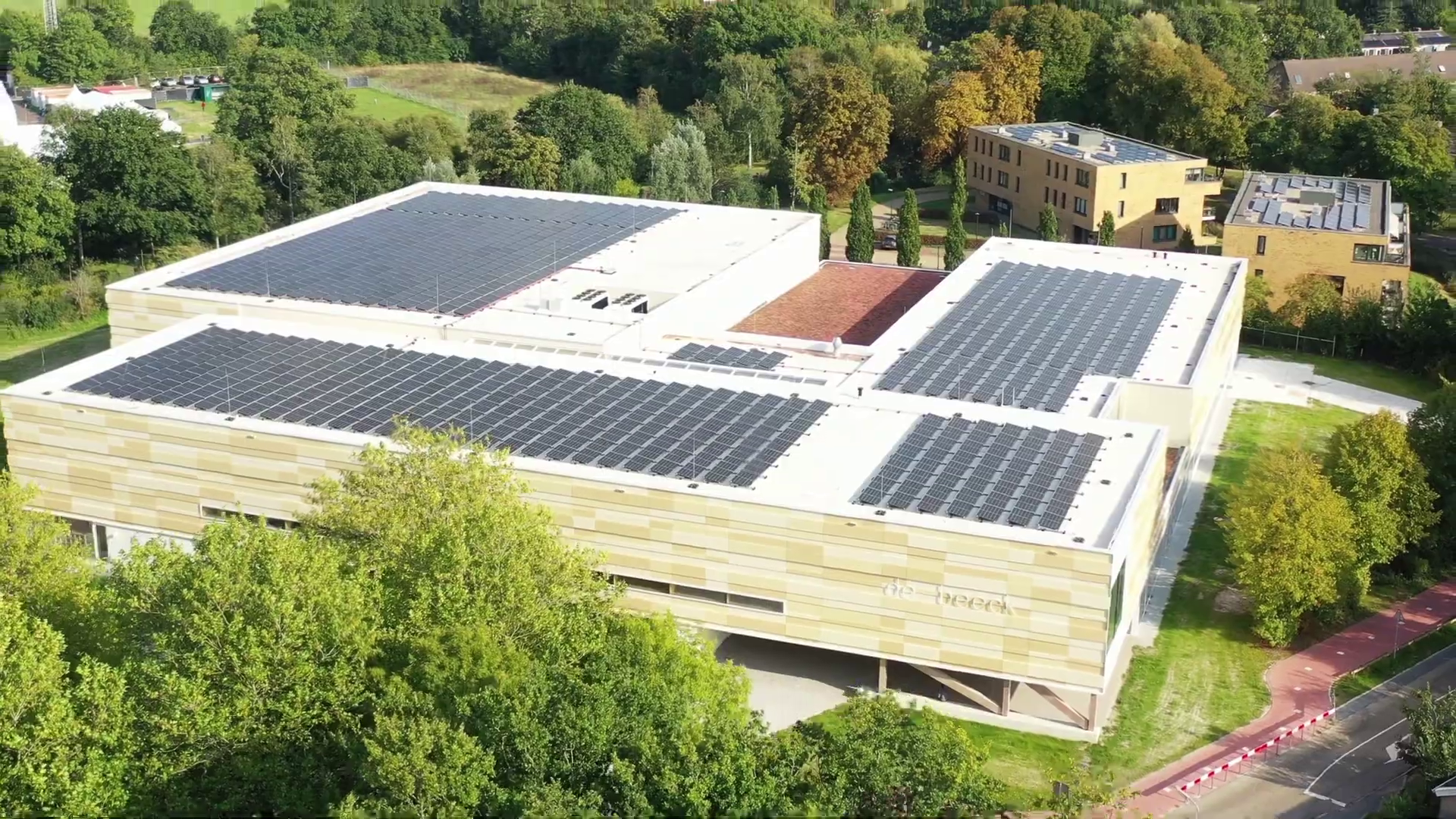 Luchtfoto van een groot gebouw met zonnepanelen op het dak, omgeven door groen en naastgelegen wegen.