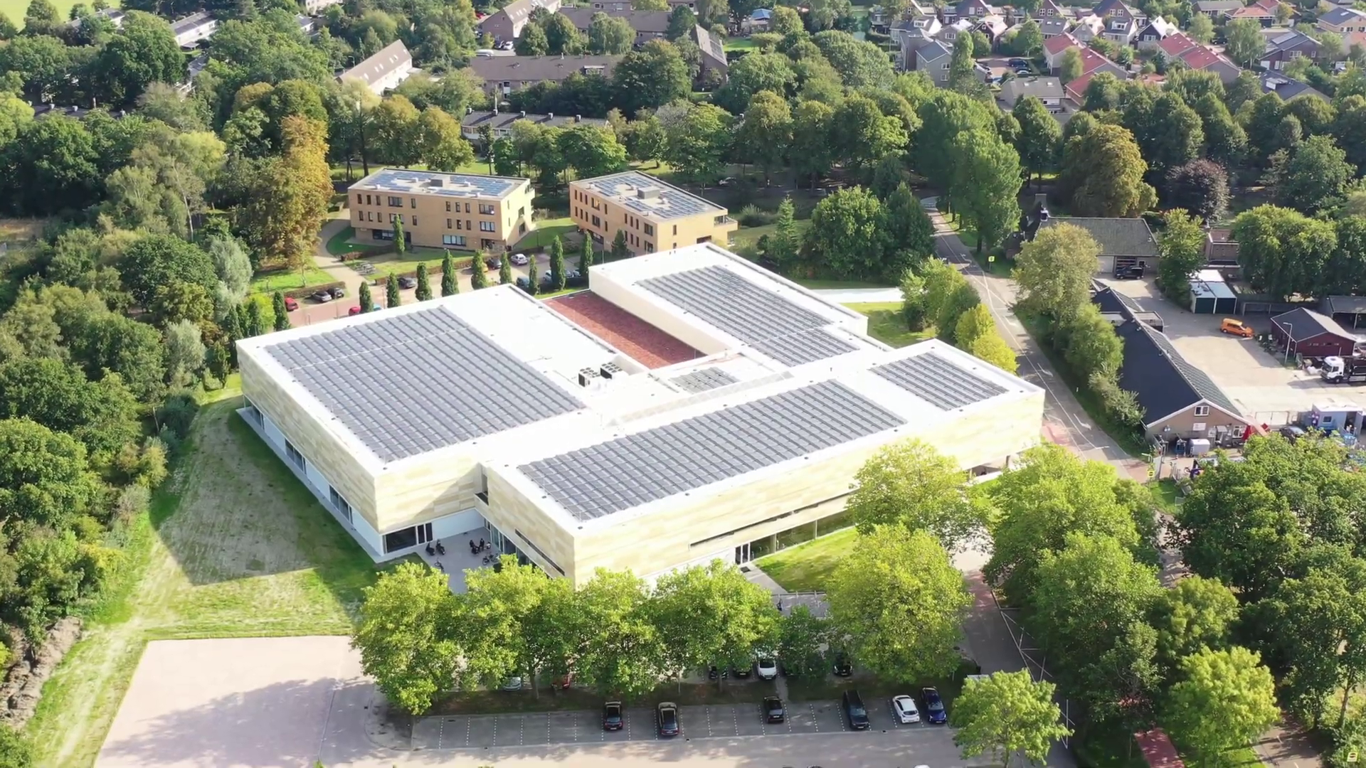 Luchtfoto van een groot, modern gebouw met zonnepanelen op het dak, omgeven door groen en nabijgelegen woningen.