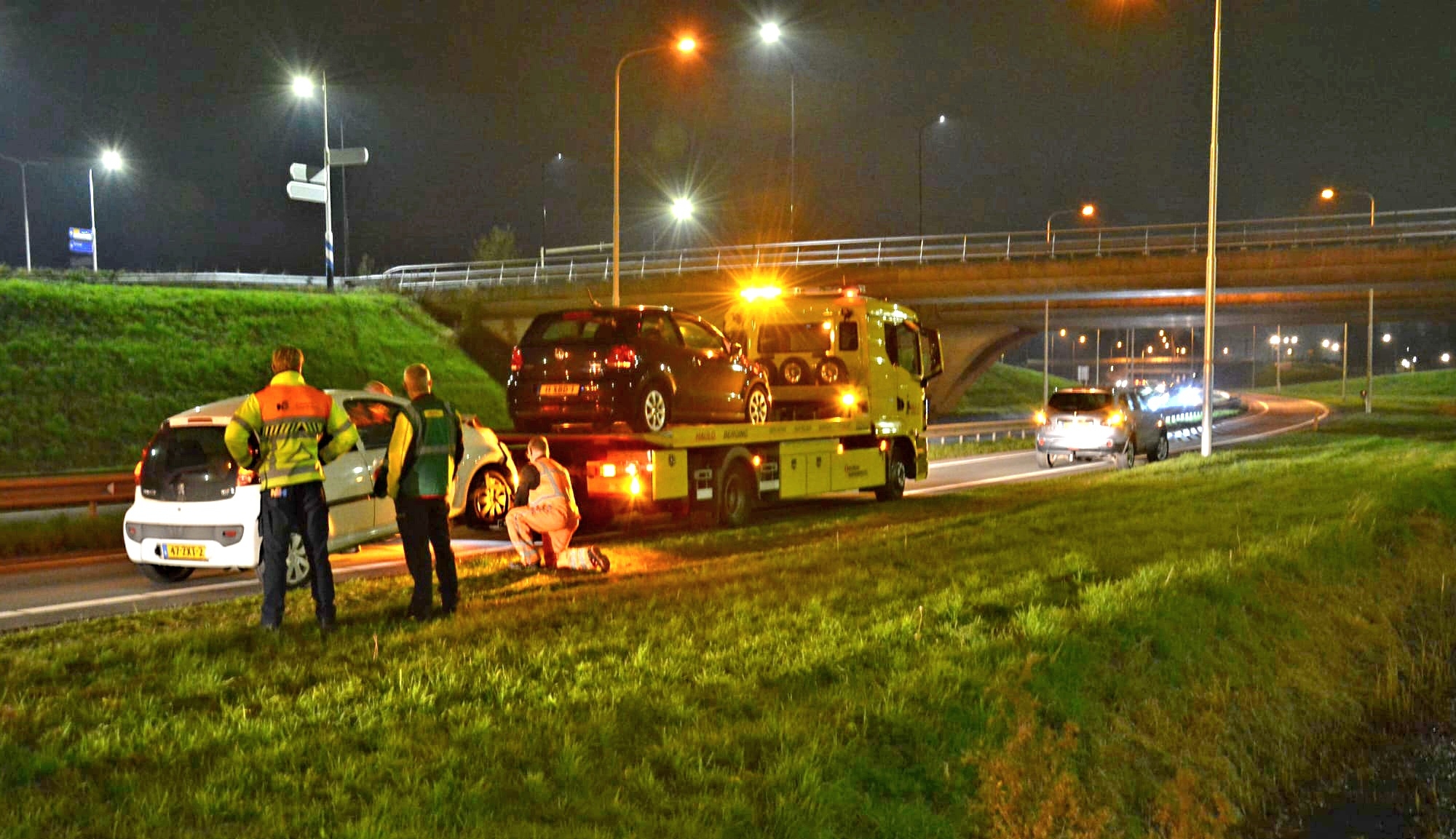 Een bergingsvoertuig met waarschuwingslichten staat stil op de snelweg met een zwarte auto op de laadbak; mensen staan naast een witte auto ernaast.