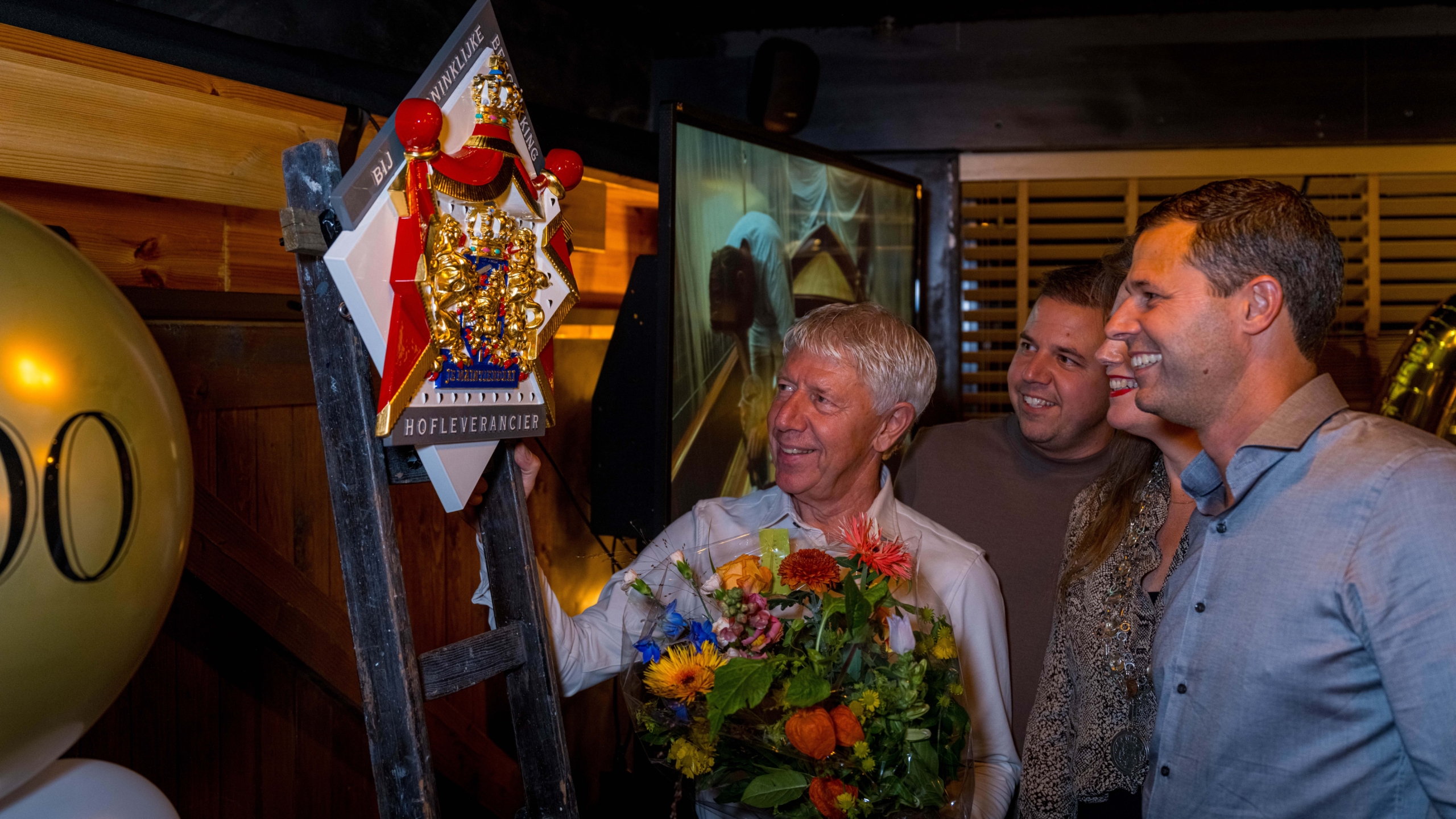 Man met bloemen wordt gefeliciteerd door drie mensen voor een schild met opschrift "Hofleverancier".