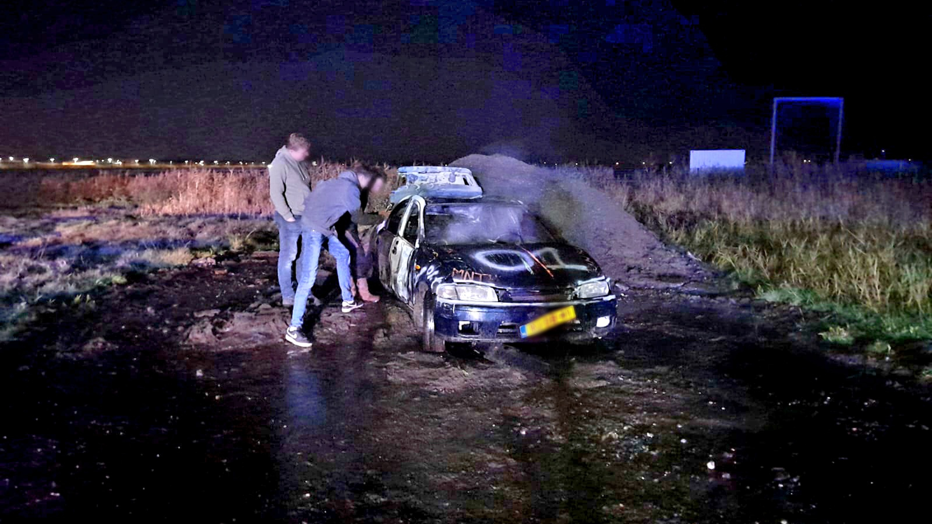 Twee personen bij een beschadigde auto in een open veld 's nachts.