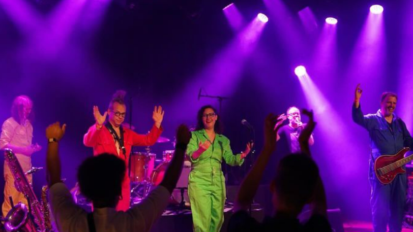 Muzikanten op het podium in kleurrijke outfits met paarse podiumverlichting, omgeven door een enthousiast publiek.