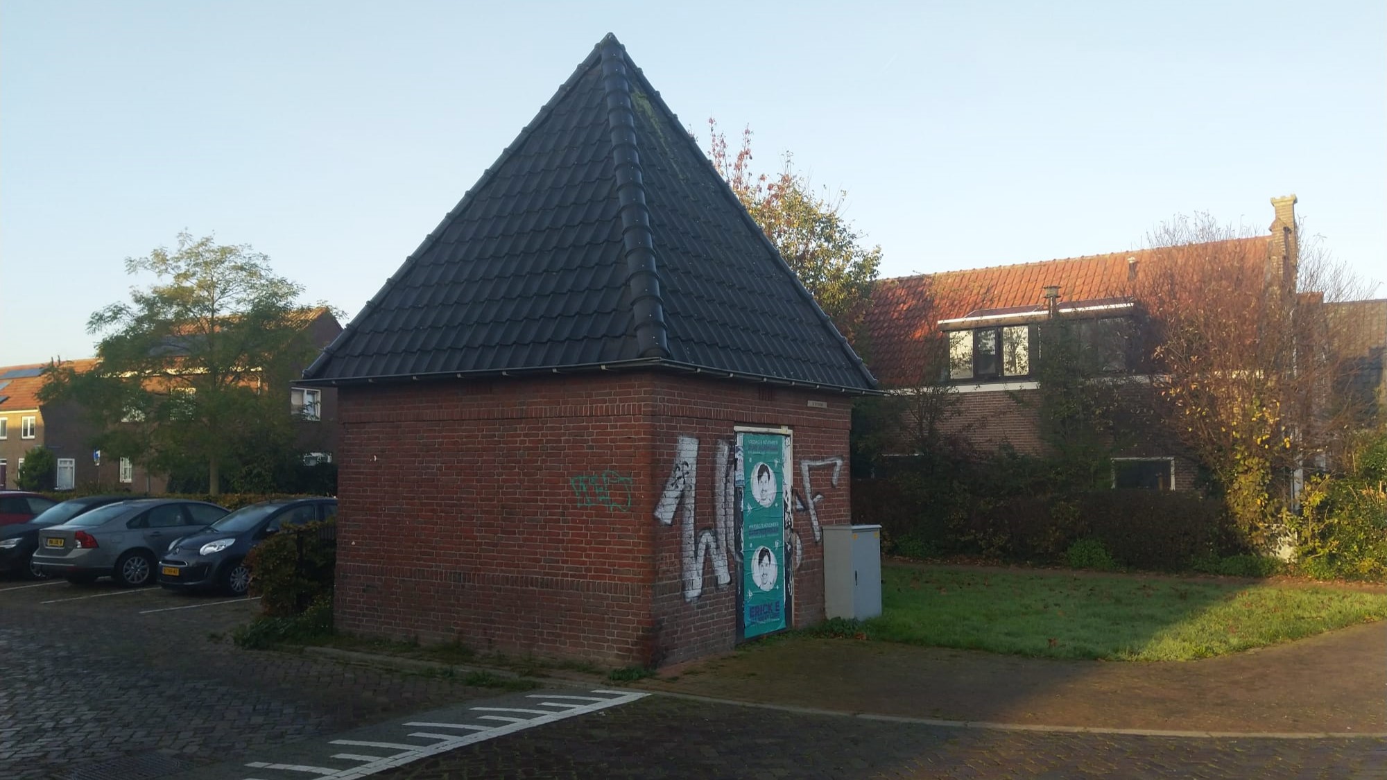 Bakstenen gebouw met piramidevormig dak, posters en graffiti op de muur, omgeven door huizen en geparkeerde auto's op een zonnige dag.