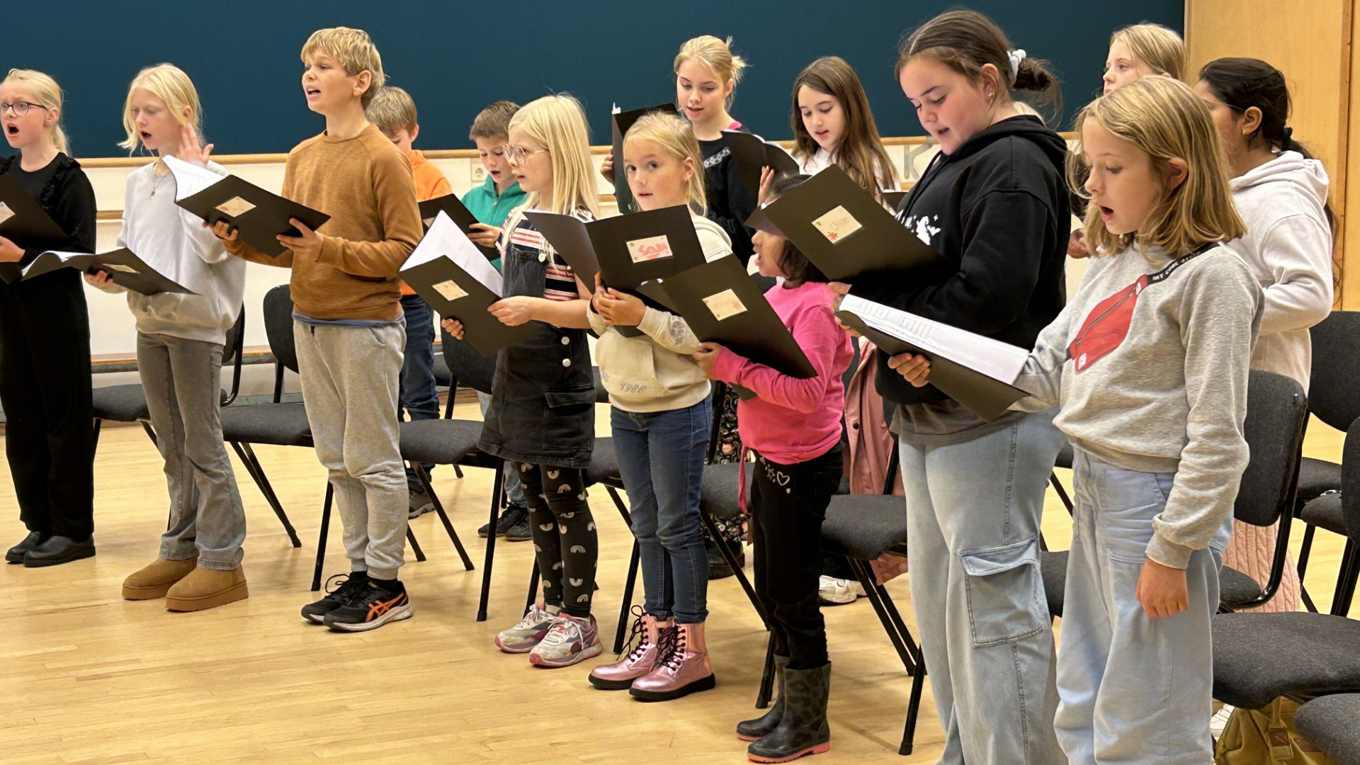 Kinderen staan in een rij en zingen uit bladmuziek terwijl ze in een repetitieruimte staan.