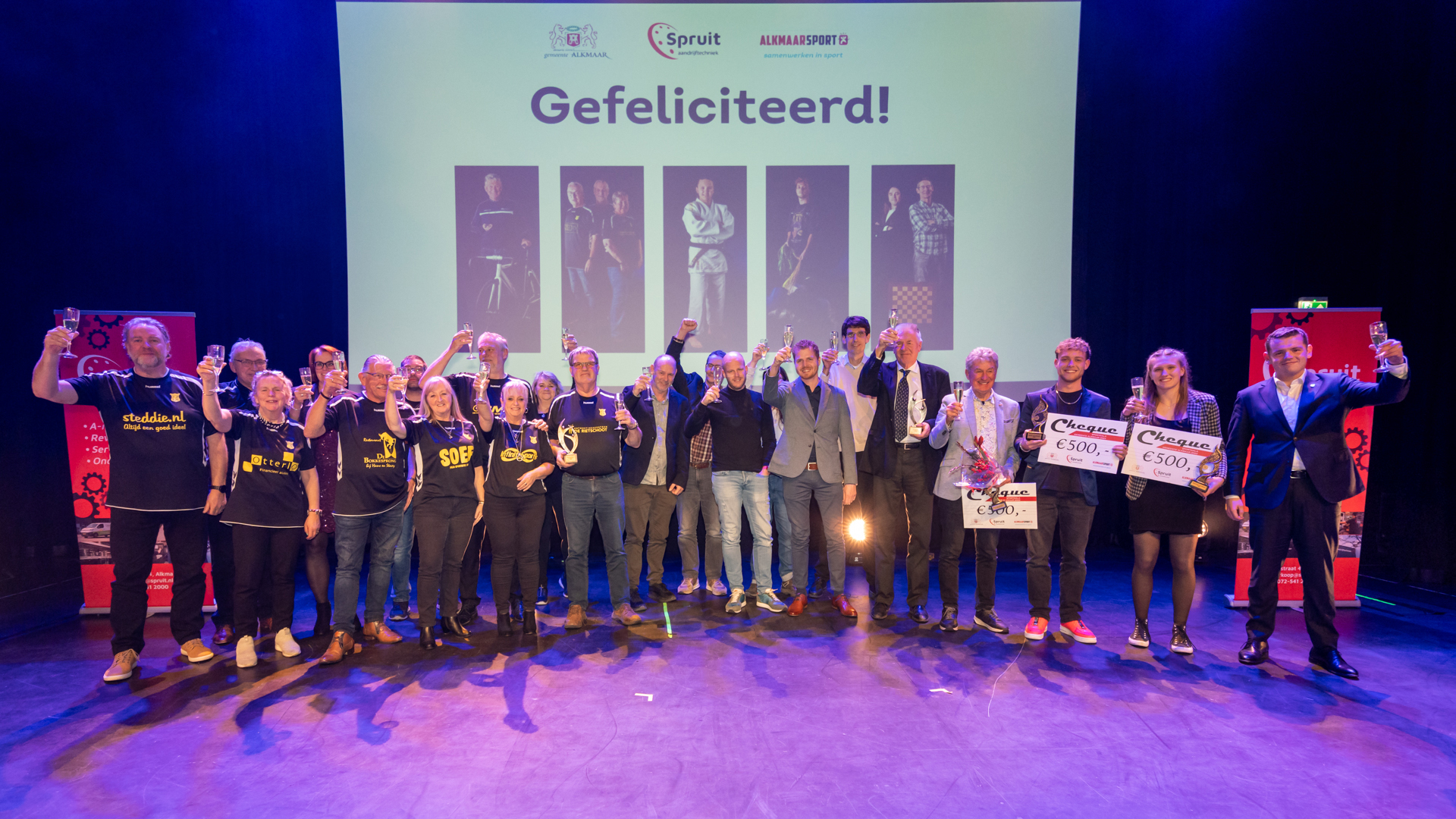 Groepsfoto van mensen op een podium met champagneglazen en cheques, met op de achtergrond een scherm met de tekst "Gefeliciteerd!".