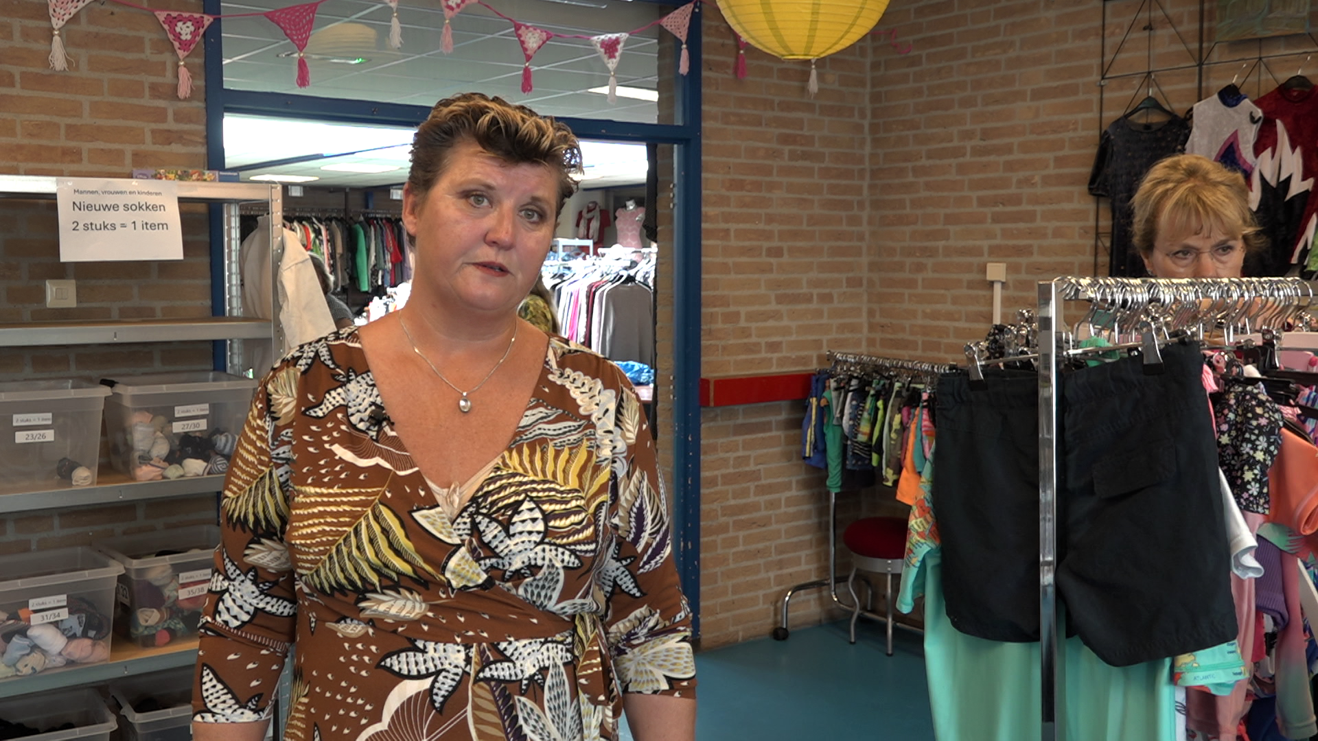 Vrouw in kringloopwinkel met kledingrekken en bord met tekst "Nieuwe sokken 2 stuks = 1 item" op de achtergrond.