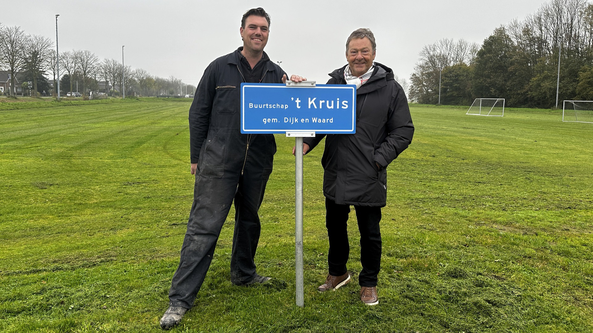 Twee mannen staan naast een blauw plaatsnaambord van buurtschap 't Kruis, gem. Dijk en Waard, op een groene grasveld met doelpalen in de achtergrond.