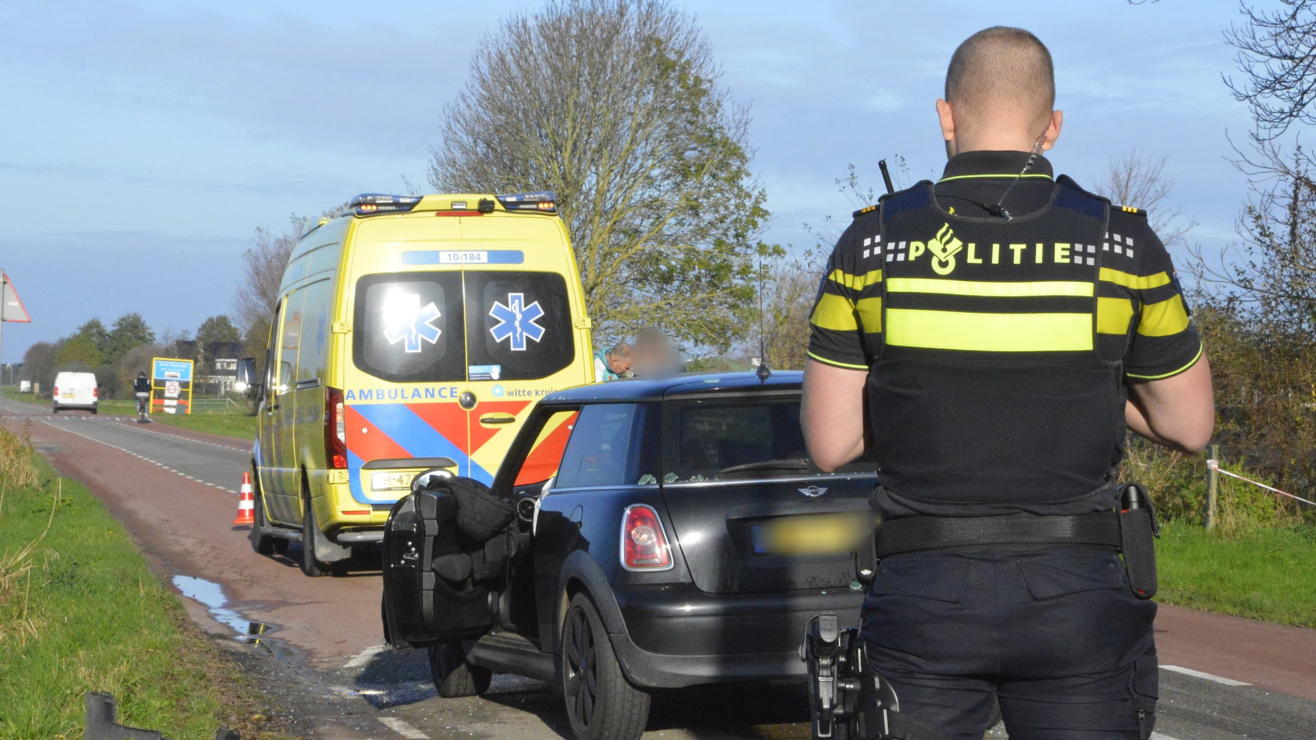 Politieagent bij zwarte auto en ambulance op een landweg.