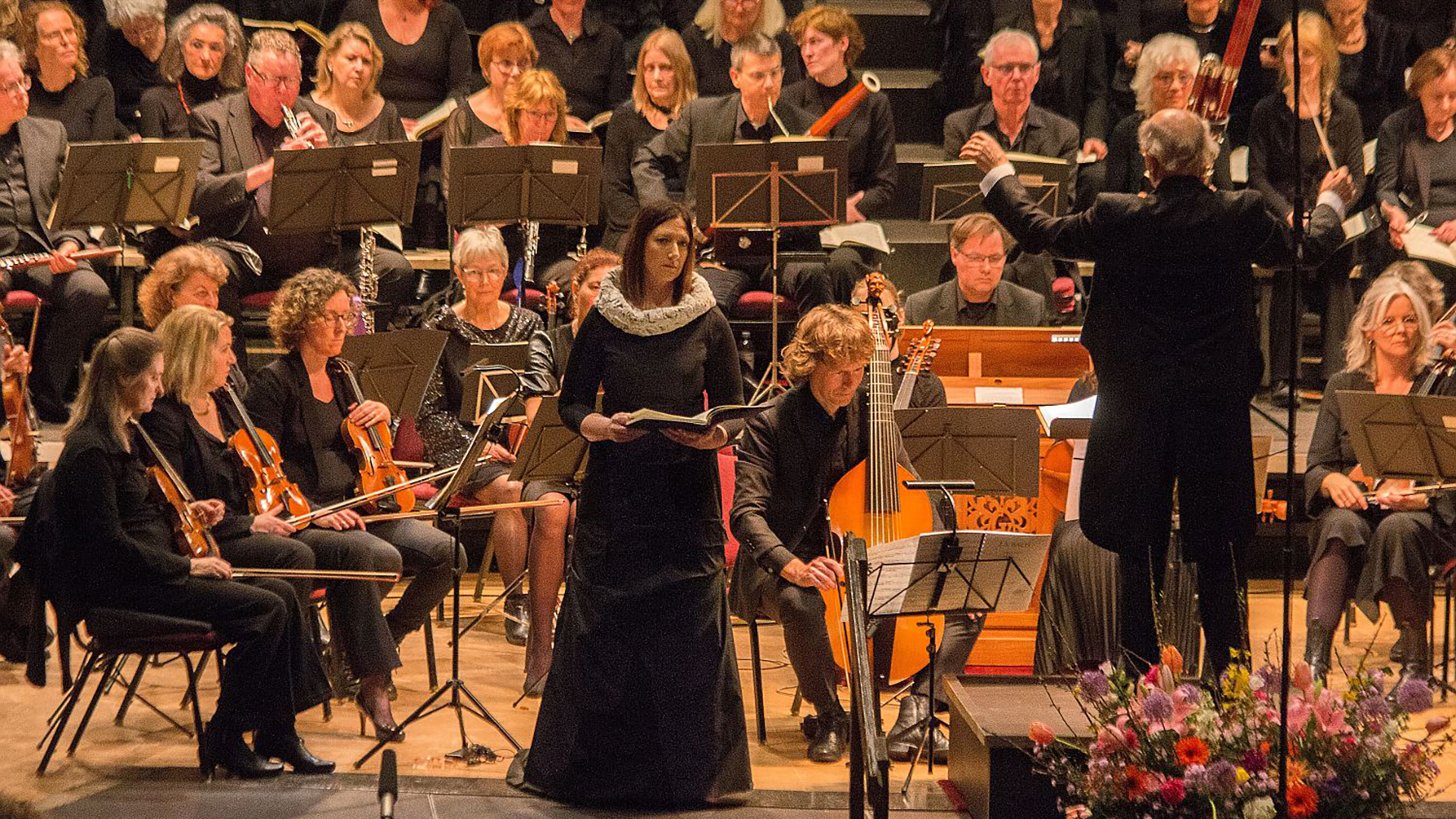 Een orkest en koor optreden met dirigent en soliste op het podium, bloemen op de voorgrond.