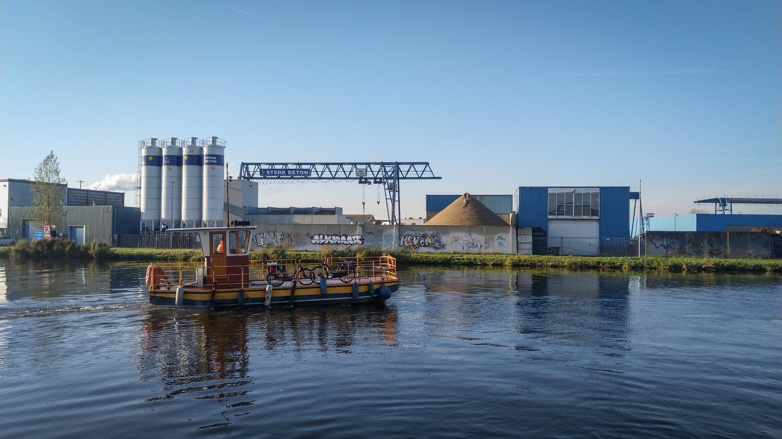 Industrieel gebied aan het water met een kleine pont in de voorgrond en gebouwen, silo’s en graffiti op de achtergrond.