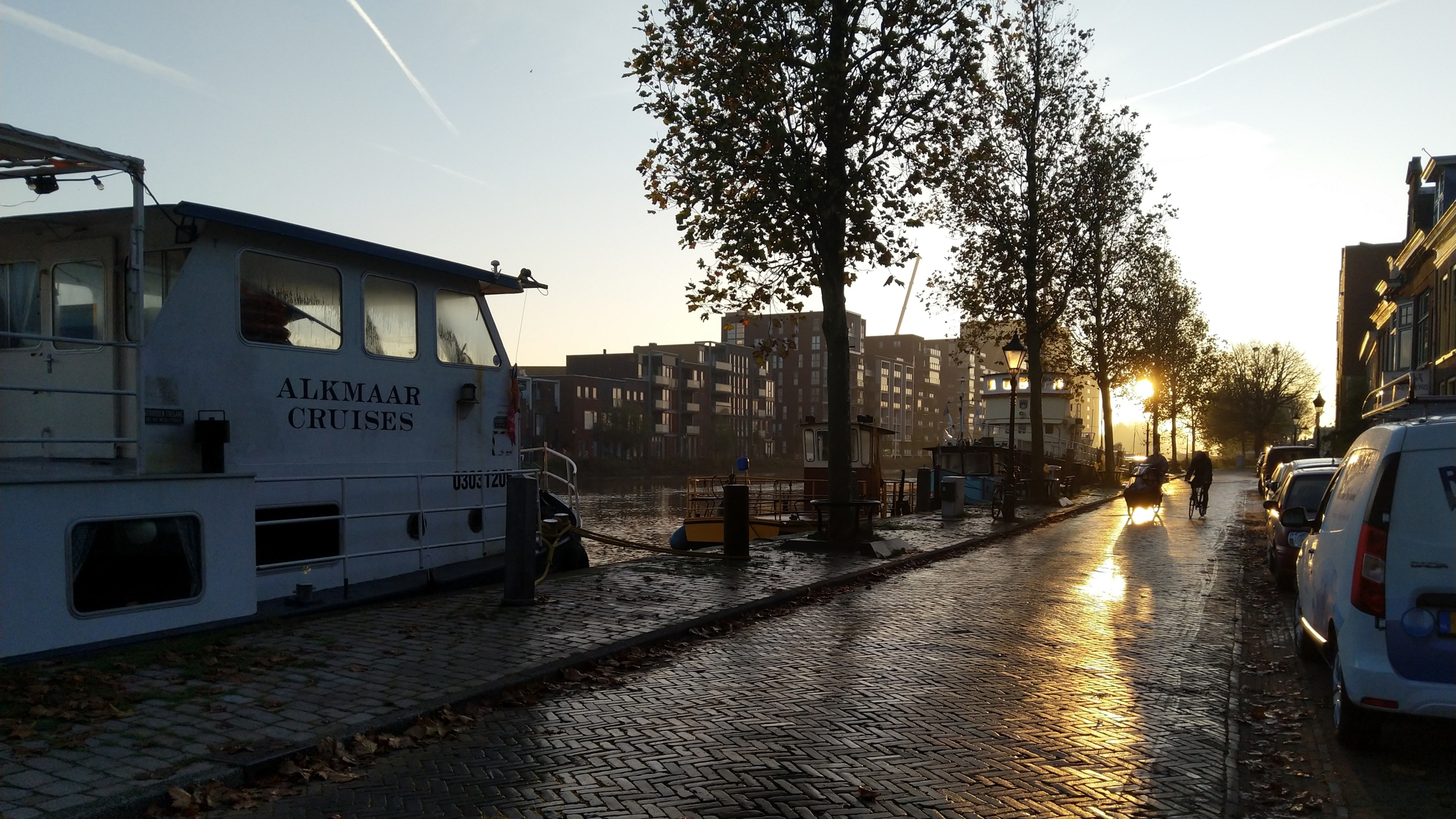 Een straat langs een kanaal met een cruiseschip aangemeerd, bomen, fietsen en auto's, bij zonsopgang.