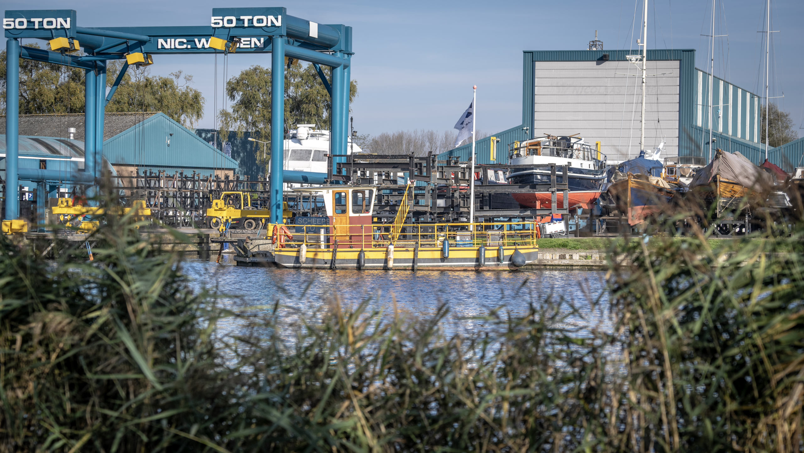 Scheepswerf met hijskraan, boten en water, omgeven door riet.